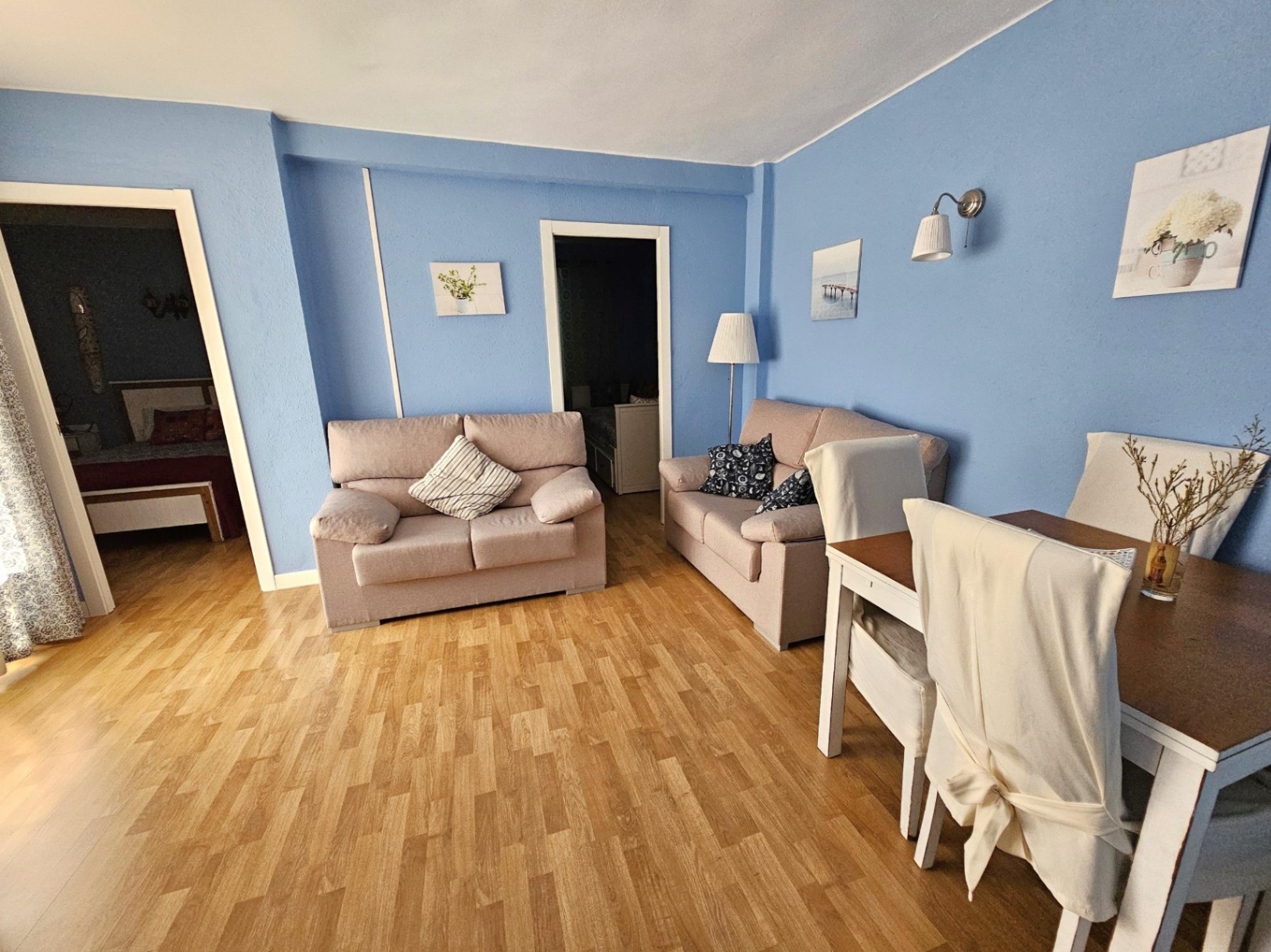 Salon-salle à manger aux murs bleus et sol en parquet. L'espace comprend deux canapés beiges et une table à manger, avec une vue dégagée sur les chambres attenantes.