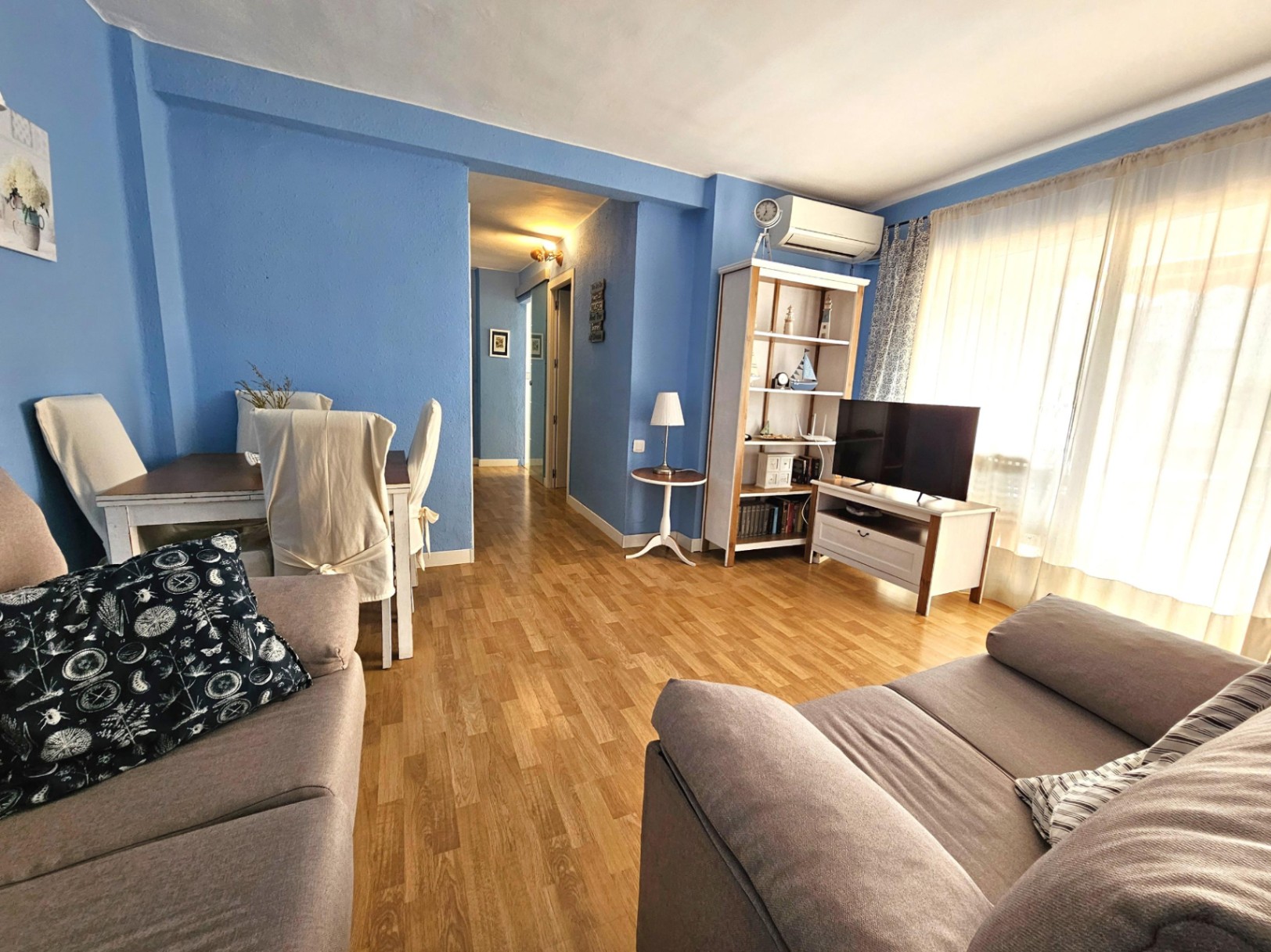 Salon-salle à manger avec murs bleus, parquet au sol et climatisation réversible. Espace de vie lumineux avec coin repas et accès au couloir.