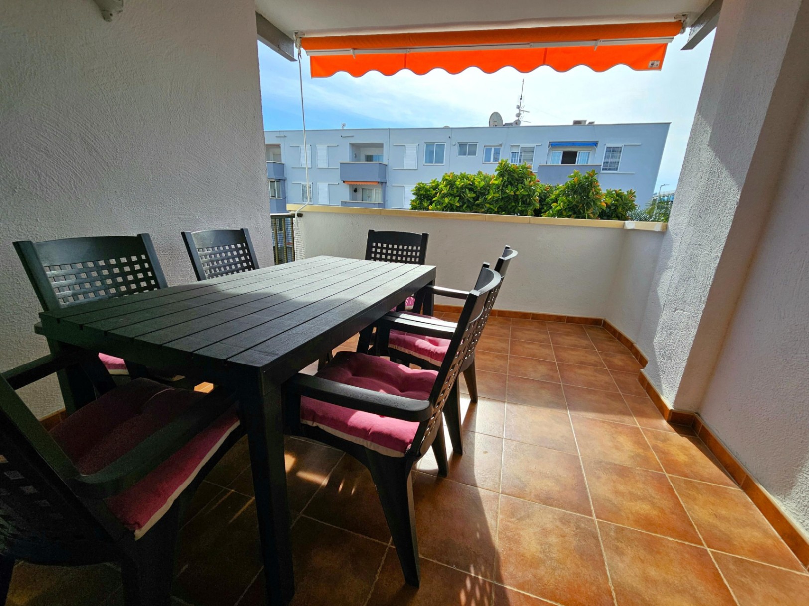 Terrasse extérieure avec carrelage en terre cuite, ensemble de repas sombre pour six personnes avec coussins roses et store banne orange rétractable.