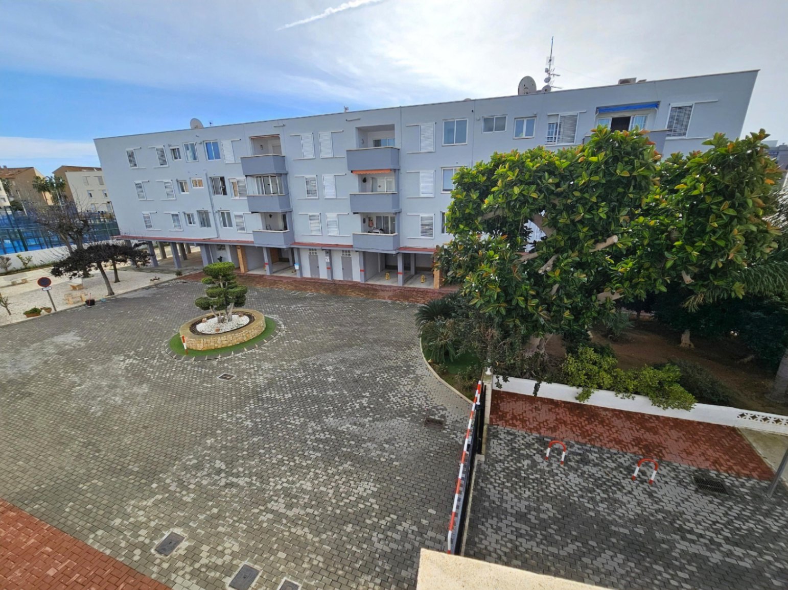 Vue extérieure du complexe résidentiel avec sa cour pavée, ses espaces verts soignés et ses emplacements de parking privés couverts sous le bâtiment.