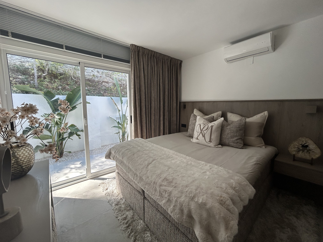 Chambre moderne avec accès direct au patio, climatisation murale et tête de lit en bois intégrée dans un design aux tons neutres.