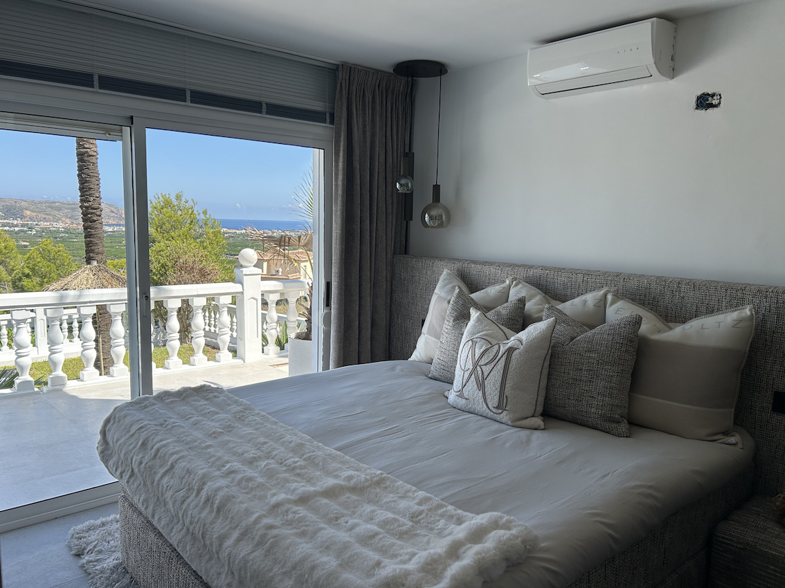 Chambre principale avec tête de lit rembourrée et climatisation, s'ouvrant sur une terrasse privée avec balustrade offrant une vue dégagée sur la mer.