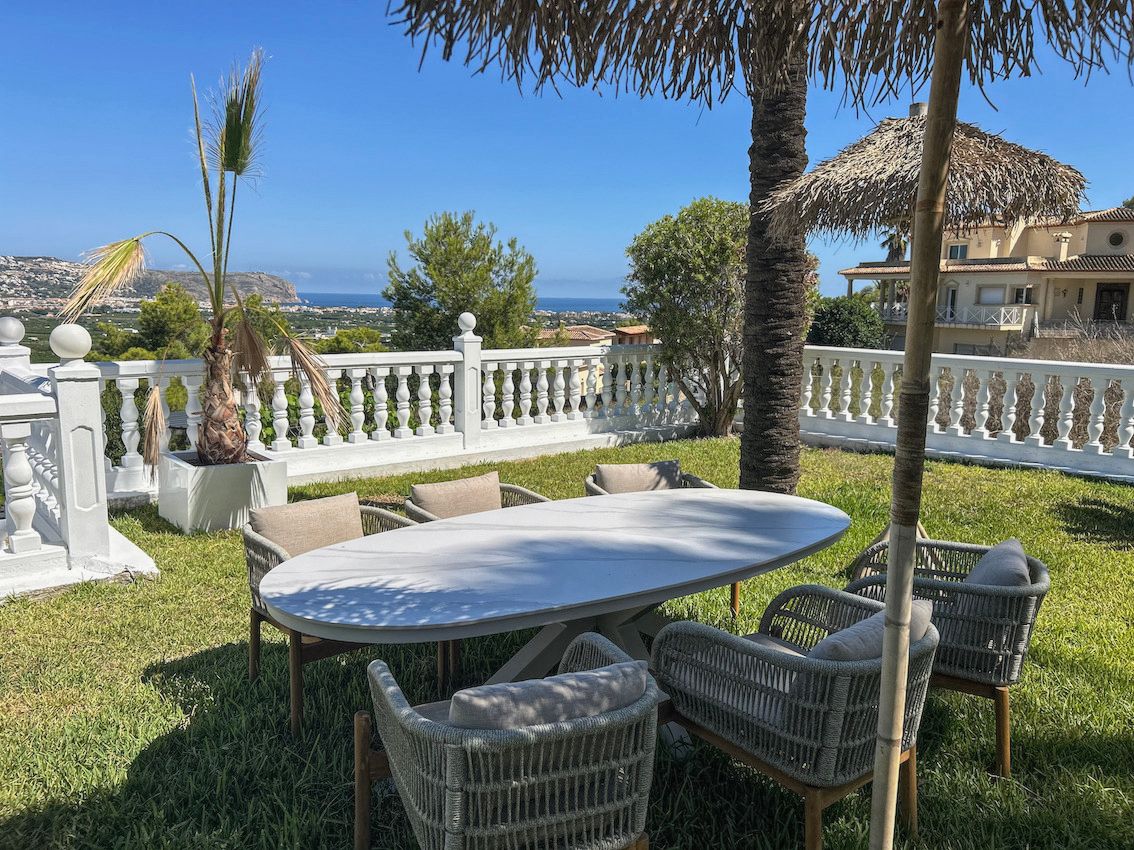 Espace repas extérieur sur pelouse avec table ovale, fauteuils en corde tressée et balustrade blanche offrant une vue dégagée sur la mer.