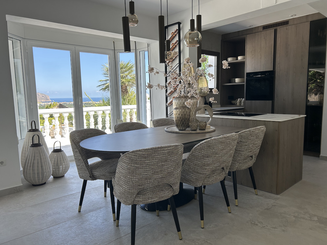 Salle à manger à aire ouverte avec table ovale et cuisine intégrée en bois, offrant une vue panoramique sur la mer grâce à de grandes baies vitrées.