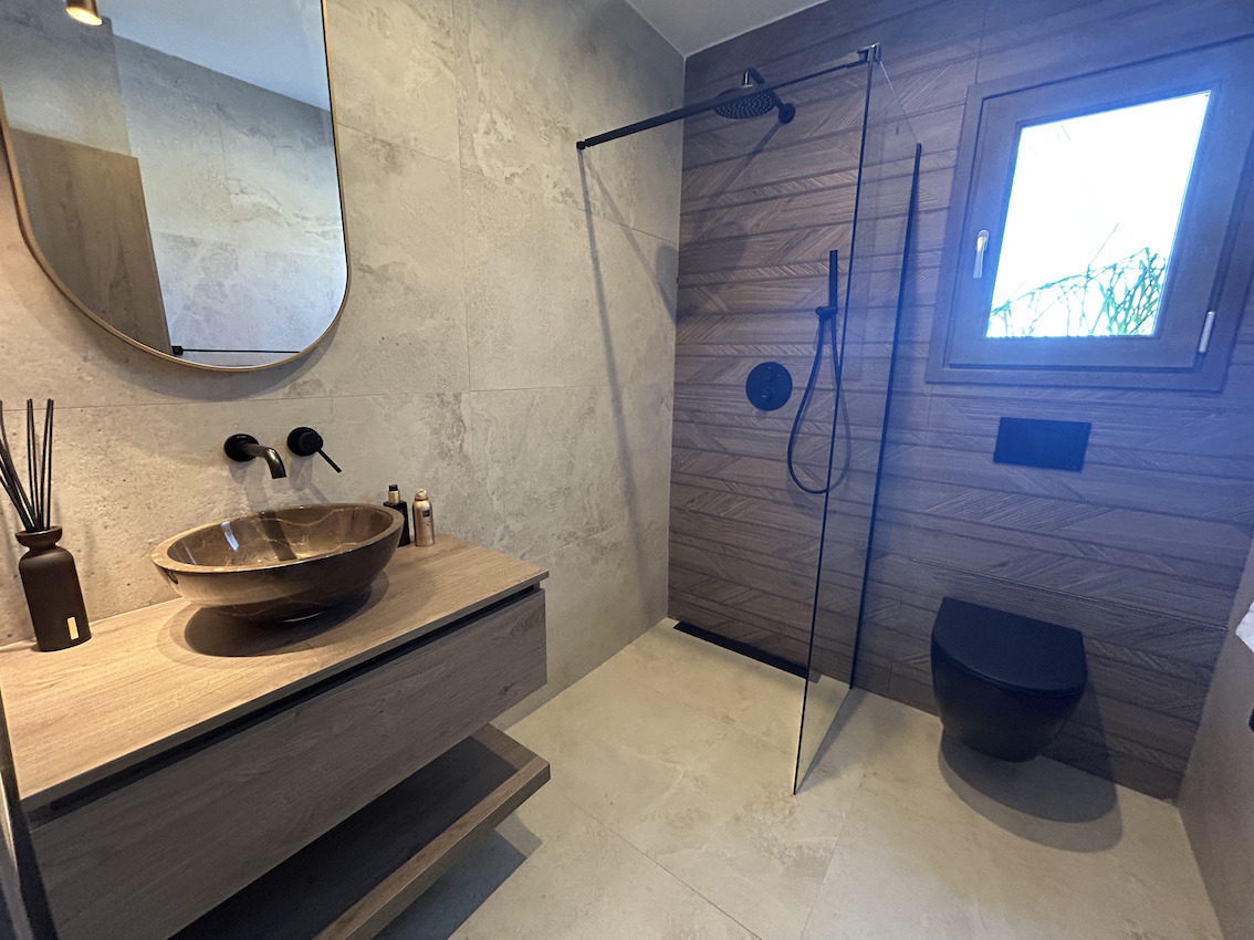 Salle de bain moderne avec douche à l'italienne, robinetterie noire, vasque en pierre sur meuble en bois suspendu et carrelage texturé.