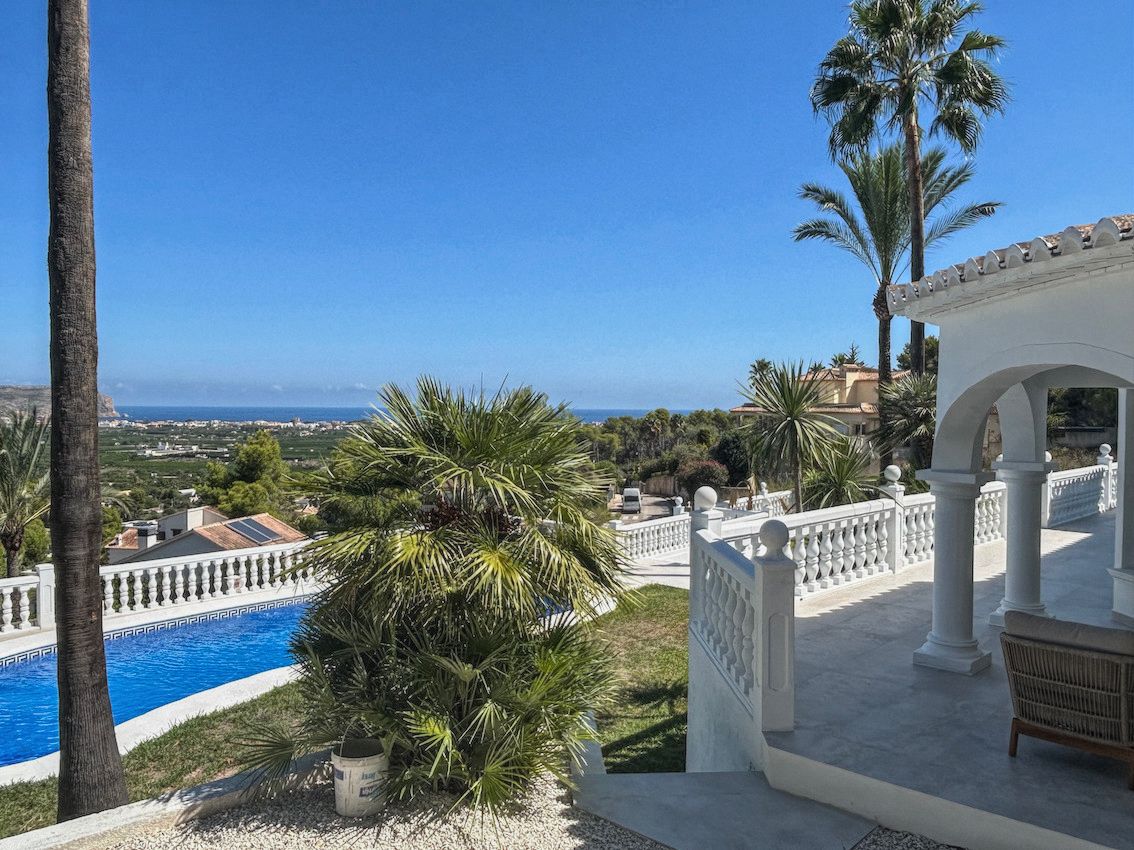 Terrasse avec balustrades blanches et porche voûté offrant une vue panoramique sur la mer et la vallée, entourée de palmiers et de la piscine.