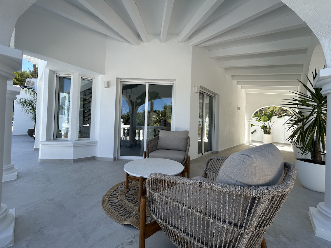 Terrasse couverte avec poutres apparentes blanches, mobilier d'extérieur moderne et sol en grès gris, offrant un espace de détente ombragé.
