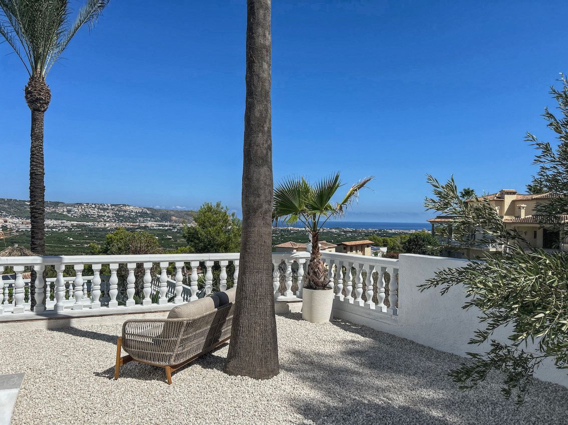 Terrasse extérieure avec sol en gravier blanc, mobilier moderne et vue panoramique sur la mer et la vallée, bordée de palmiers et d'une balustrade blanche.
