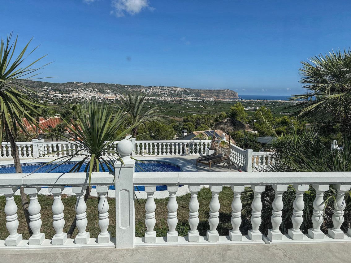 Terrasse surélevée avec balustrade blanche offrant une vue panoramique sur la vallée et la mer Méditerranée, surplombant la piscine et la végétation locale.