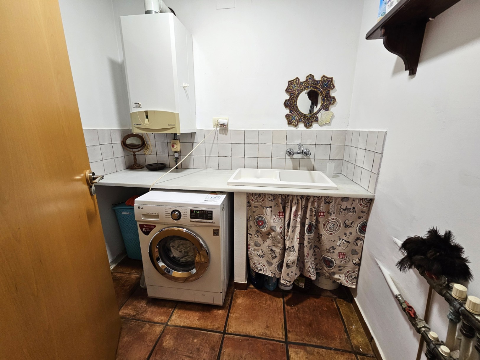 Buanderie pratique avec sol en terre cuite artisanale, équipée d'un lave-linge, d'un évier double et d'une chaudière murale sur un plan de travail blanc.