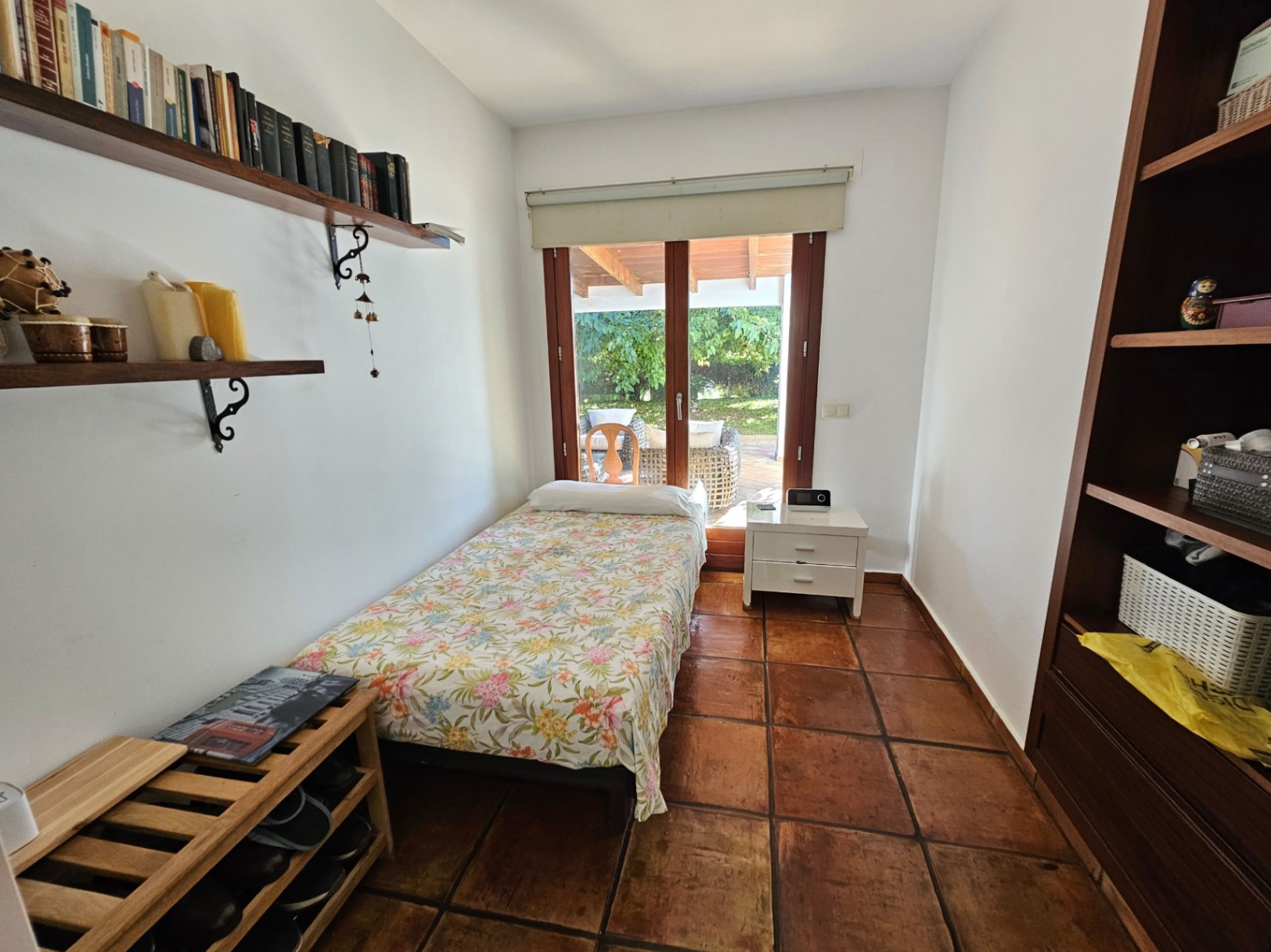 Chambre individuelle avec sols en terre cuite artisanale, étagères en bois et accès direct à la terrasse couverte et au jardin par une grande porte vitrée.