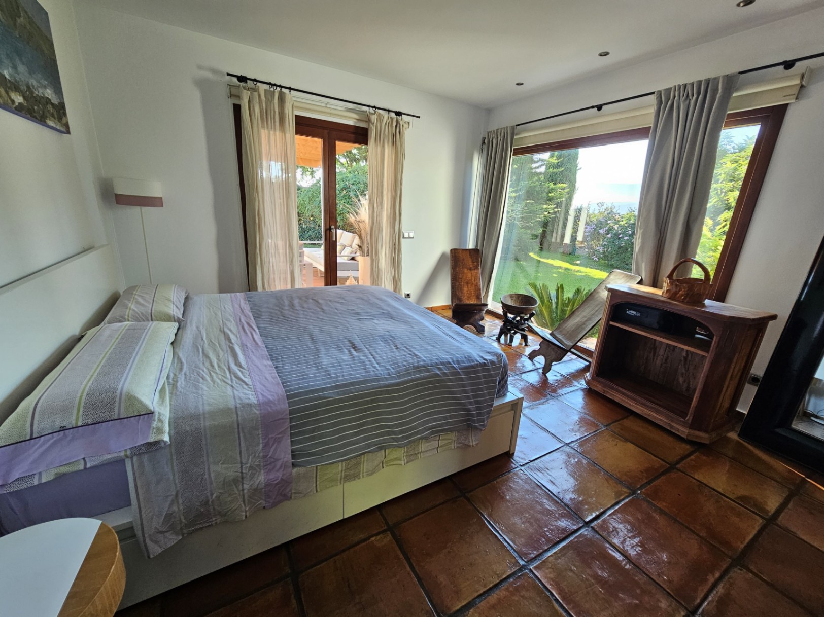 Chambre lumineuse avec sols en terre cuite artisanale, accès direct au jardin et grandes baies vitrées offrant une vue sur les espaces verts extérieurs.