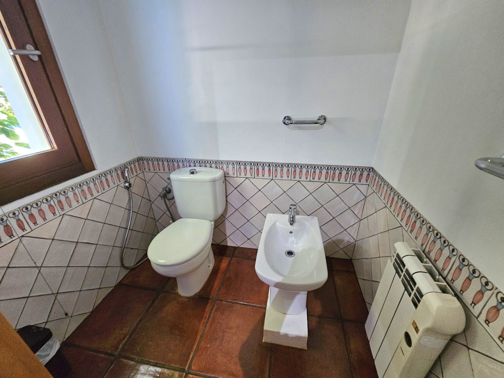 Coin toilette avec bidet, sols en terre cuite artisanale et carrelage mural orné d'une frise peinte à la main. Équipé d'un radiateur et d'une fenêtre en bois.