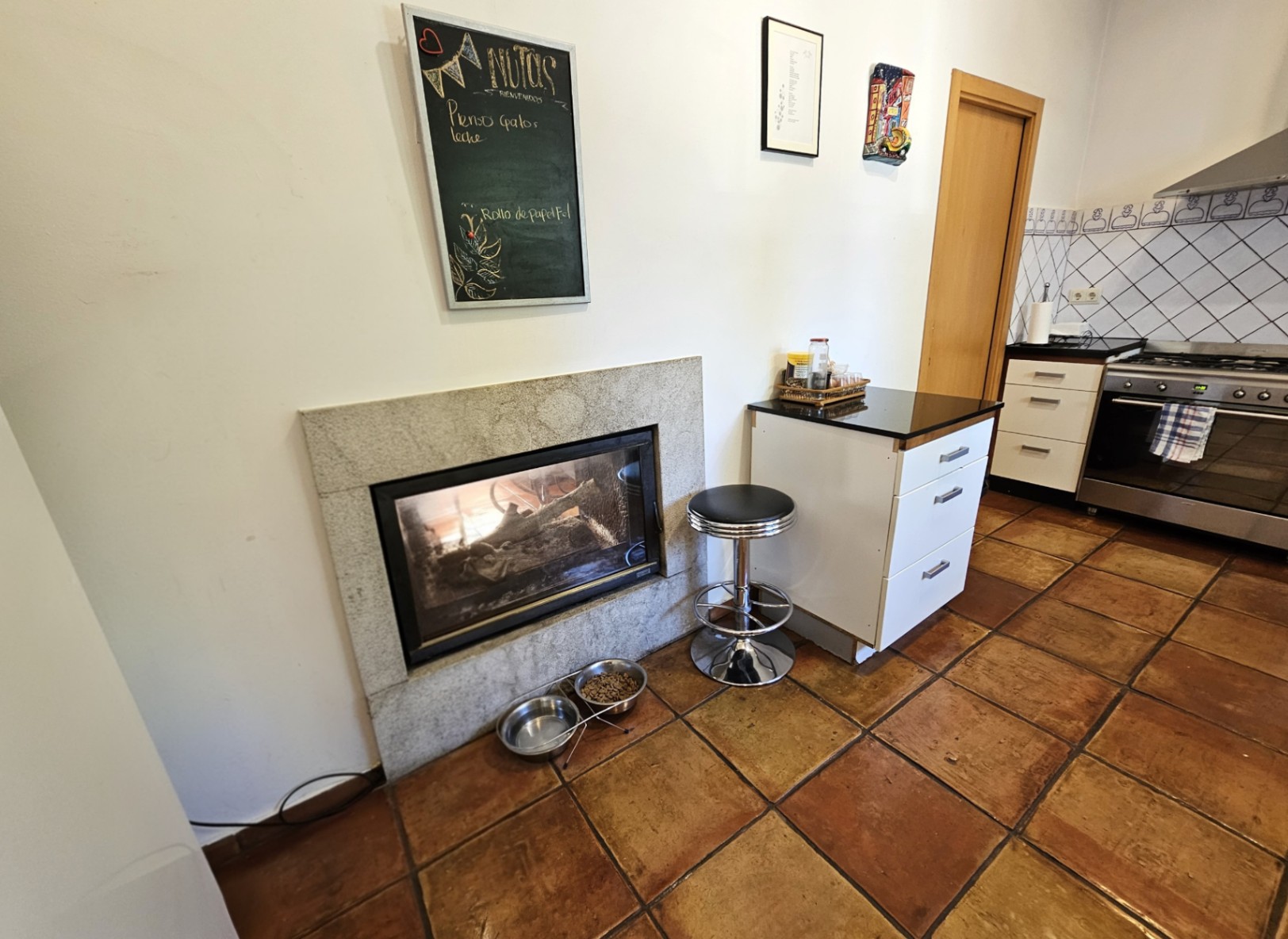 Cuisine avec sol en terre cuite artisanale et cheminée encastrée avec cadre en pierre. Comprend des meubles blancs, un plan de travail sombre et des carreaux peints à la main.