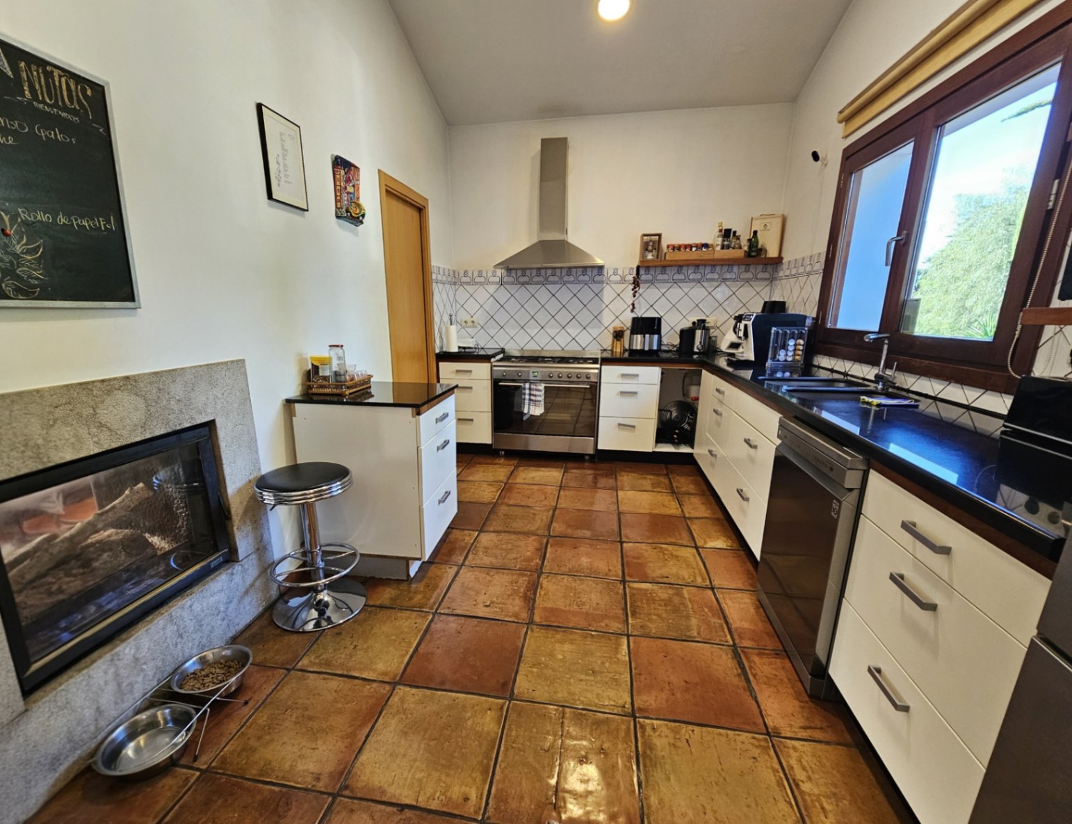 Cuisine équipée avec sol en terre cuite artisanale, plans de travail sombres et cheminée double face intégrée dans un cadre en pierre.