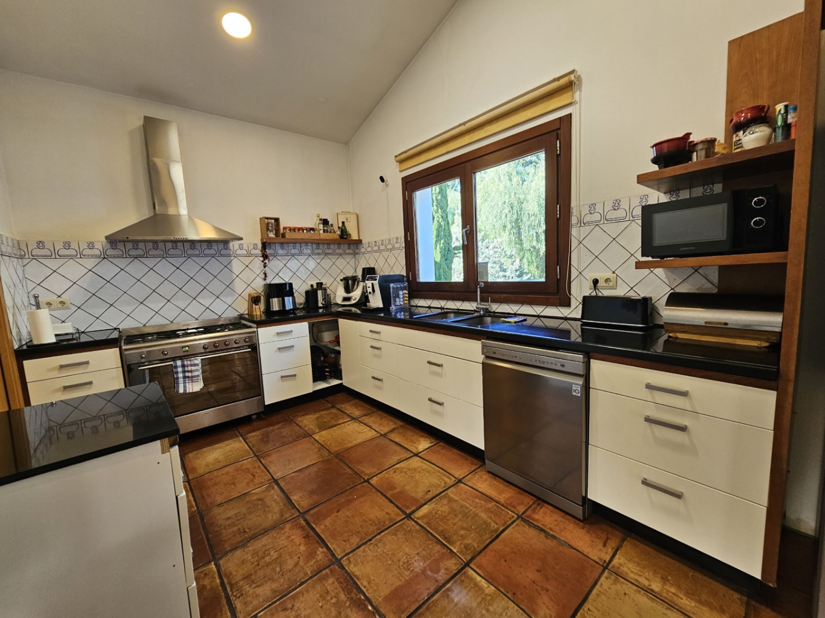 Cuisine équipée avec sols en terre cuite artisanale, plans de travail sombres et appareils en acier inoxydable, incluant une grande cuisinière à gaz et une hotte.