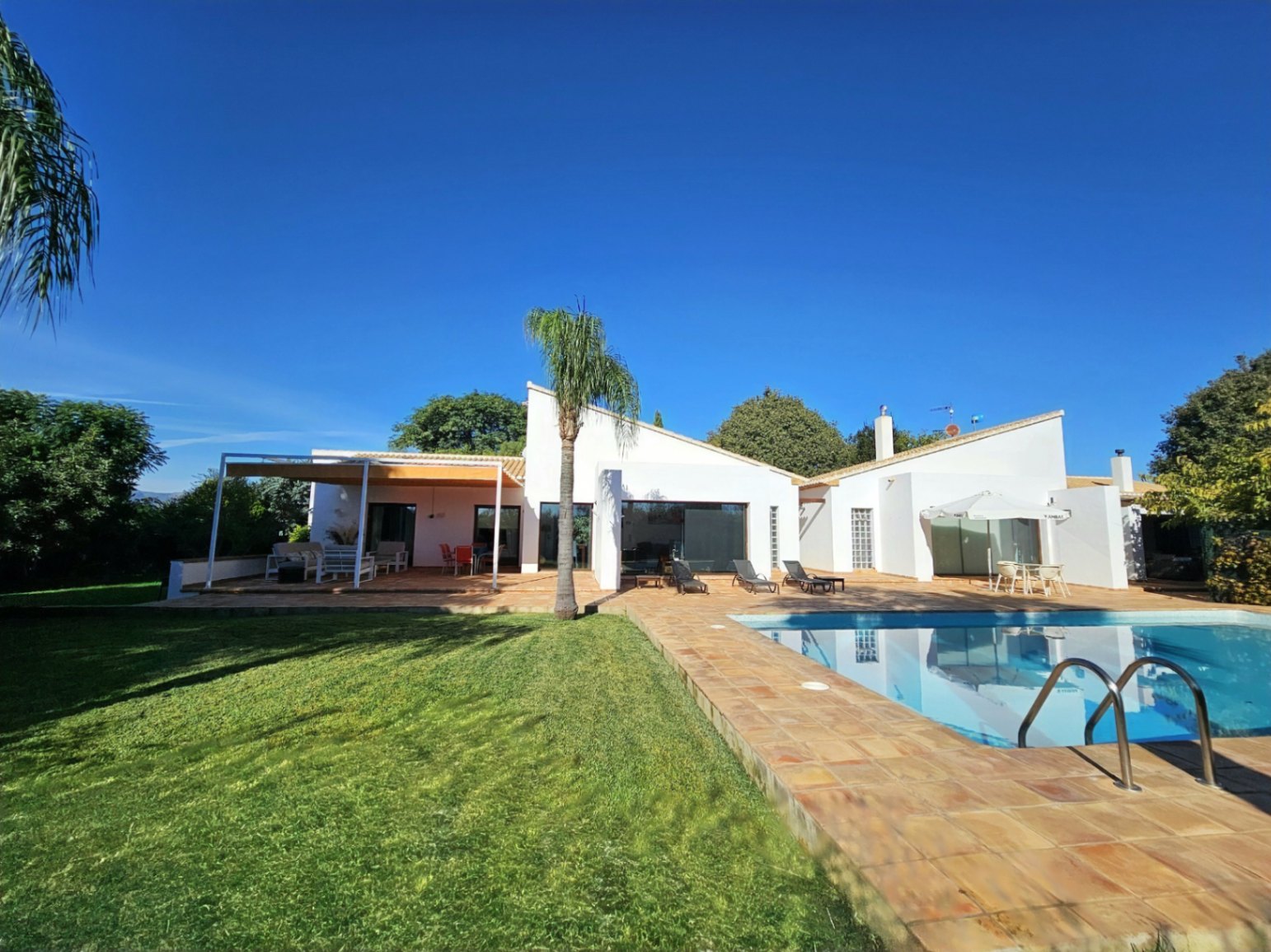 Extérieur de la villa avec piscine, terrasse en terre cuite artisanale, pelouse soignée et architecture moderne aux lignes épurées sous un ciel dégagé.