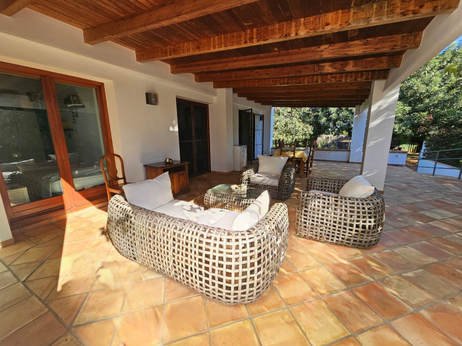Grande terrasse couverte avec poutres en bois apparentes, sol en terre cuite artisanale et salon de jardin en rotin tressé face aux extérieurs.