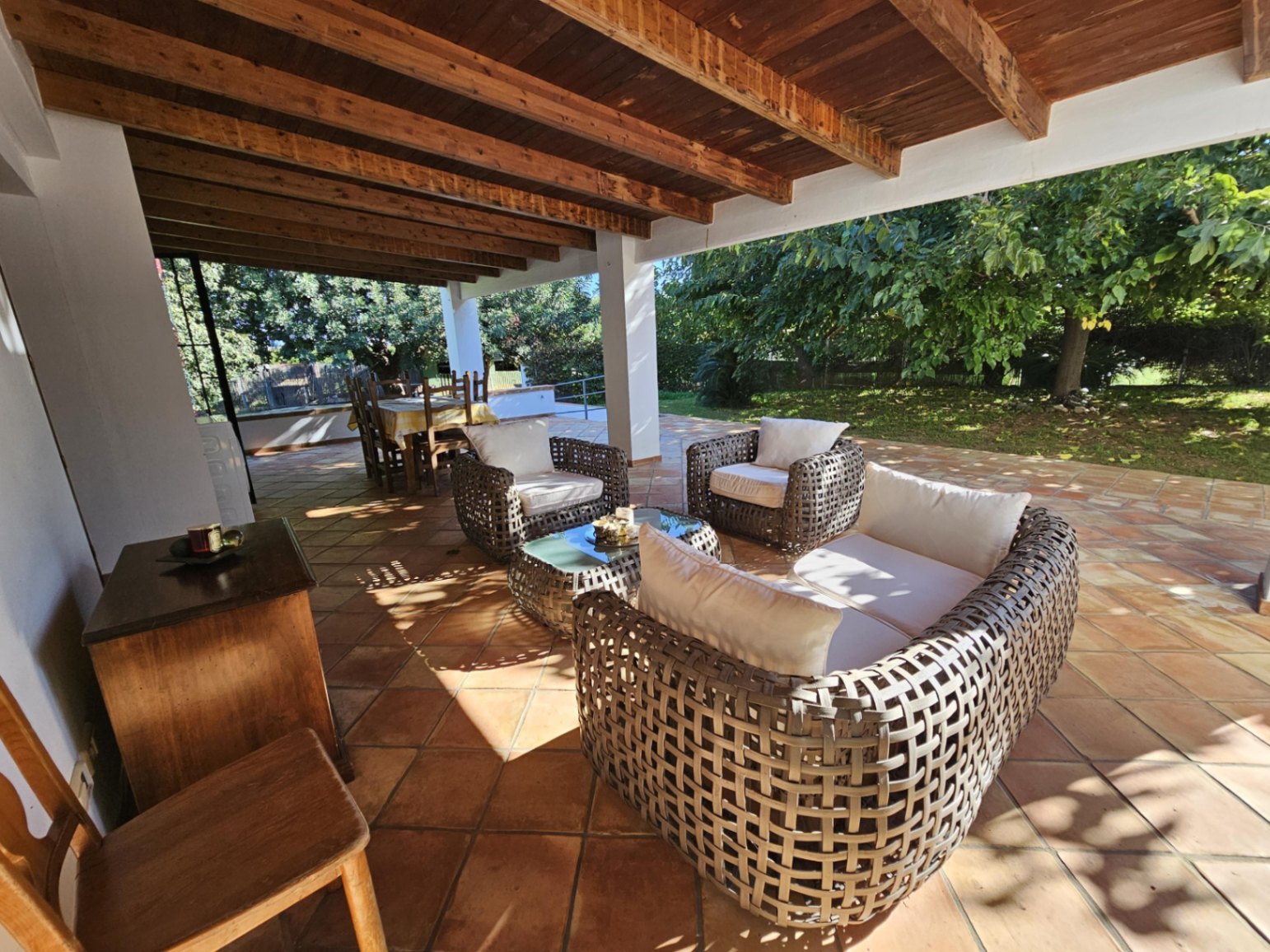 Grande terrasse couverte avec poutres en bois apparentes, sol en terre cuite artisanale et salon de jardin face au parc arboré.
