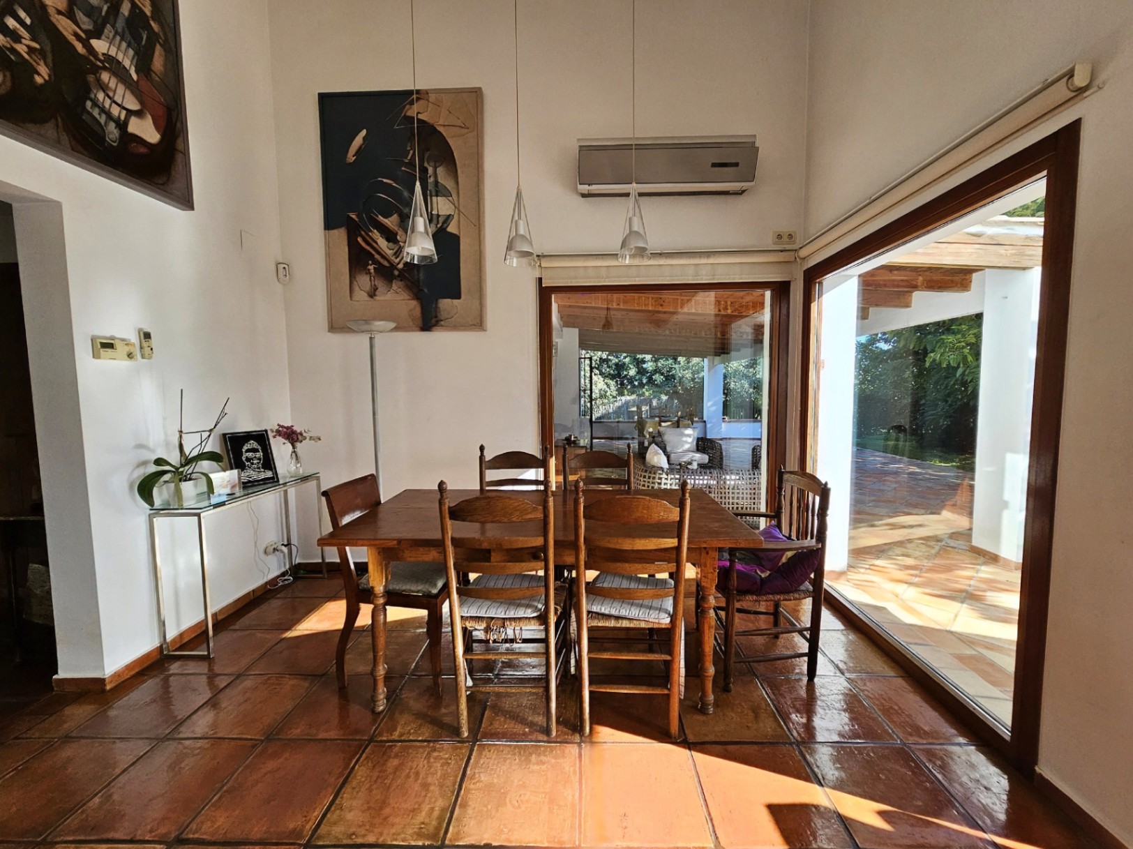 Salle à manger lumineuse avec sols en terre cuite artisanale, table en bois massif et grandes baies vitrées s'ouvrant sur la terrasse couverte et le jardin.