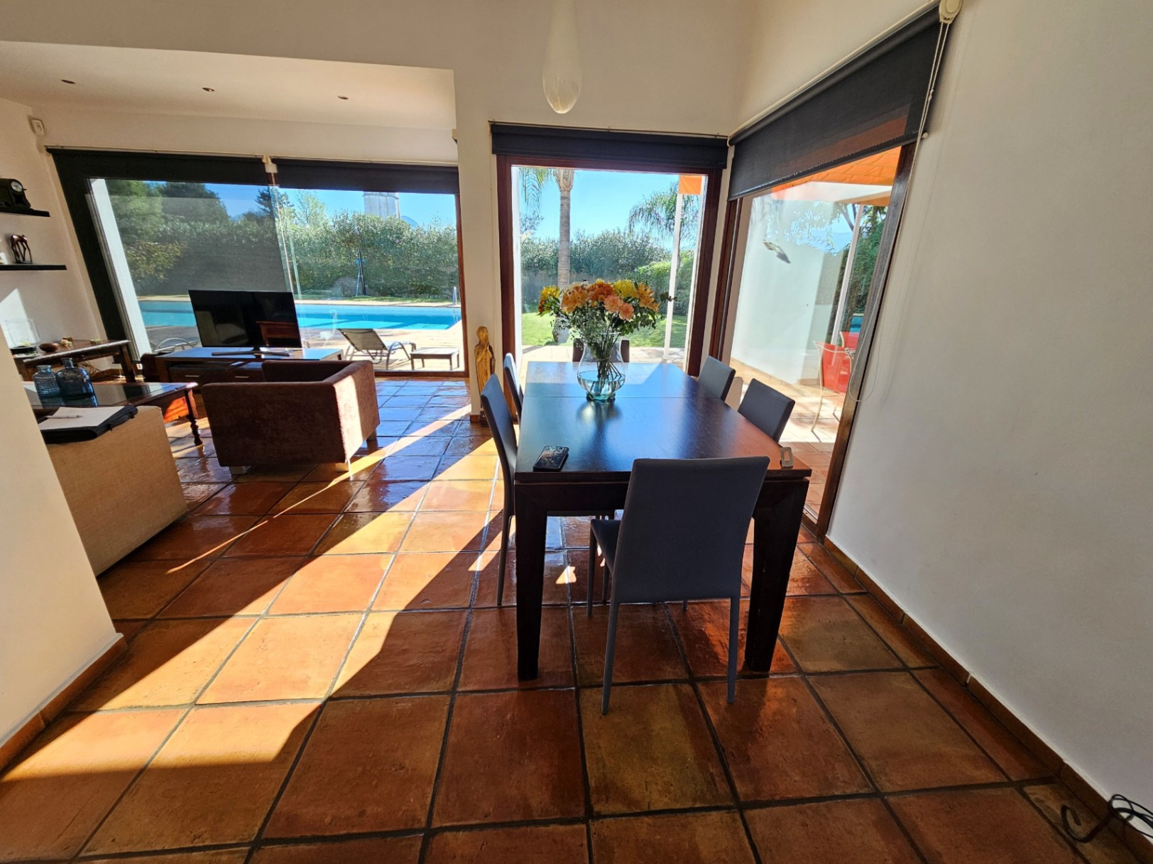 Salle à manger lumineuse avec sols en terre cuite artisanale, table en bois sombre et larges baies vitrées offrant une vue sur la piscine et le jardin.
