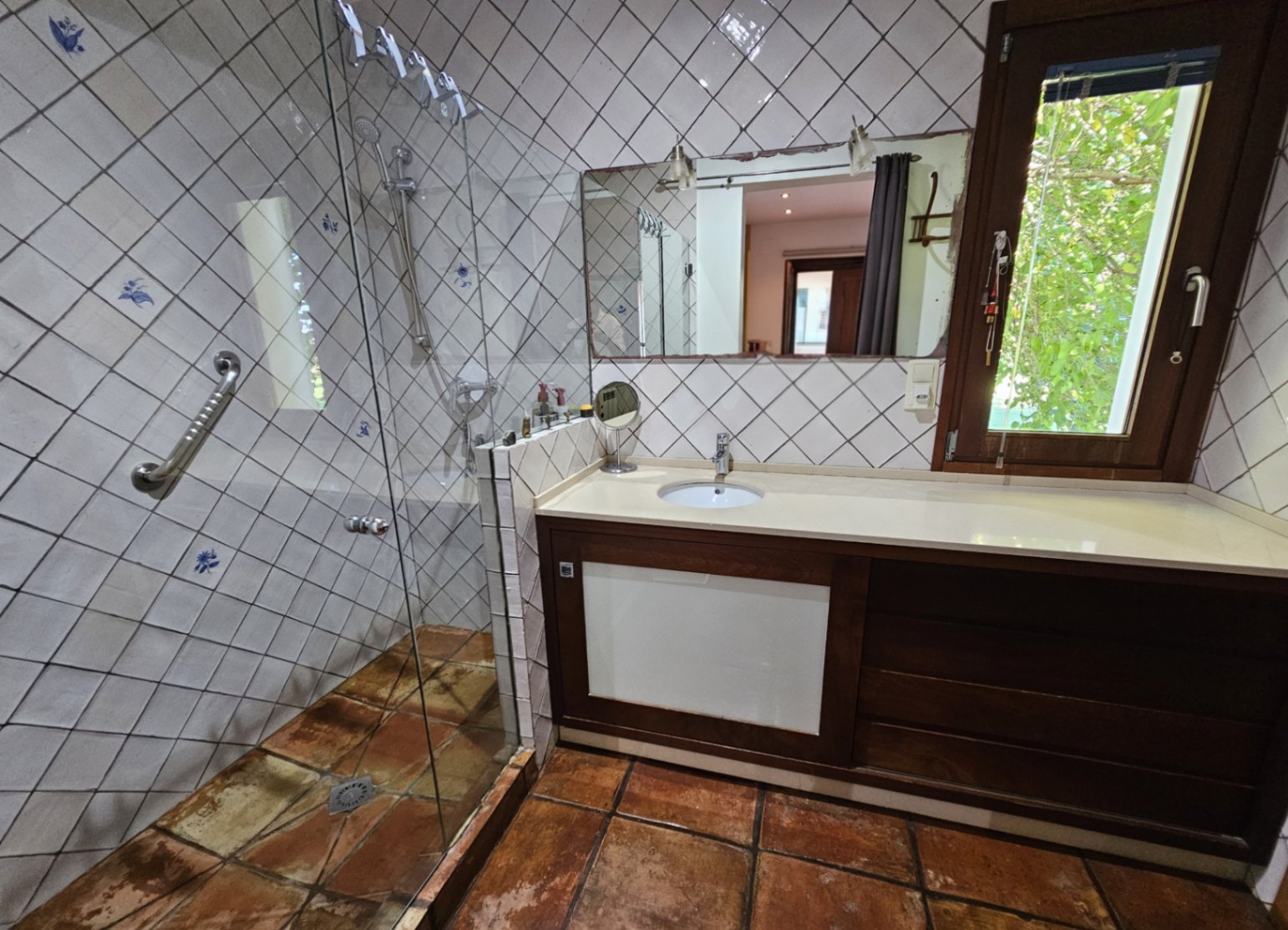 Salle de bain avec carrelage peint à la main, douche à l'italienne vitrée et sol en terre cuite. Le meuble vasque en bois est complété par une fenêtre ouvrant sur la verdure.