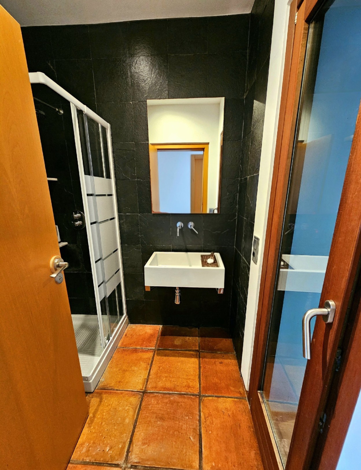 Salle de bains avec sol en terre cuite artisanale, murs en pierre noire texturée, lavabo suspendu moderne et cabine de douche vitrée.