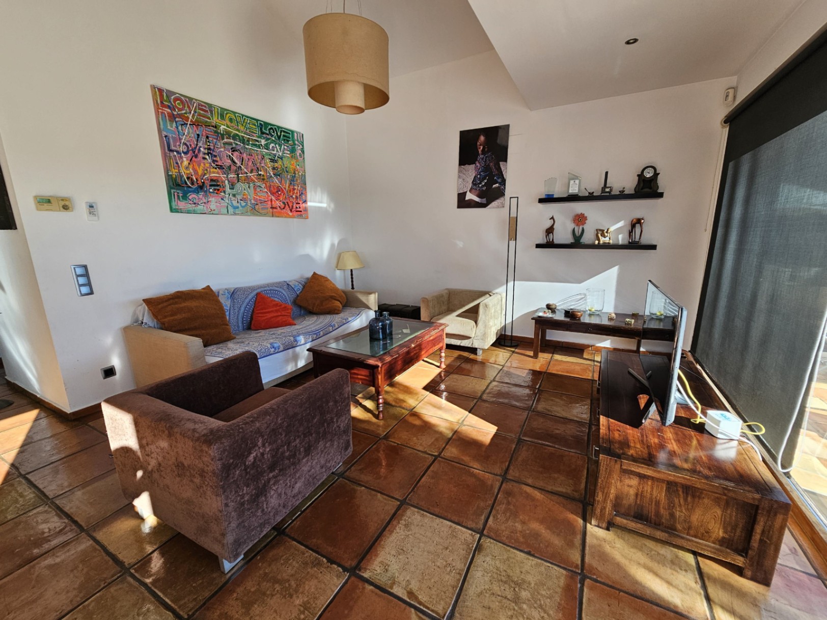 Salon avec des sols en terre cuite artisanale et du mobilier en bois. La lumière naturelle met en valeur les œuvres d'art et l'atmosphère chaleureuse de la pièce.