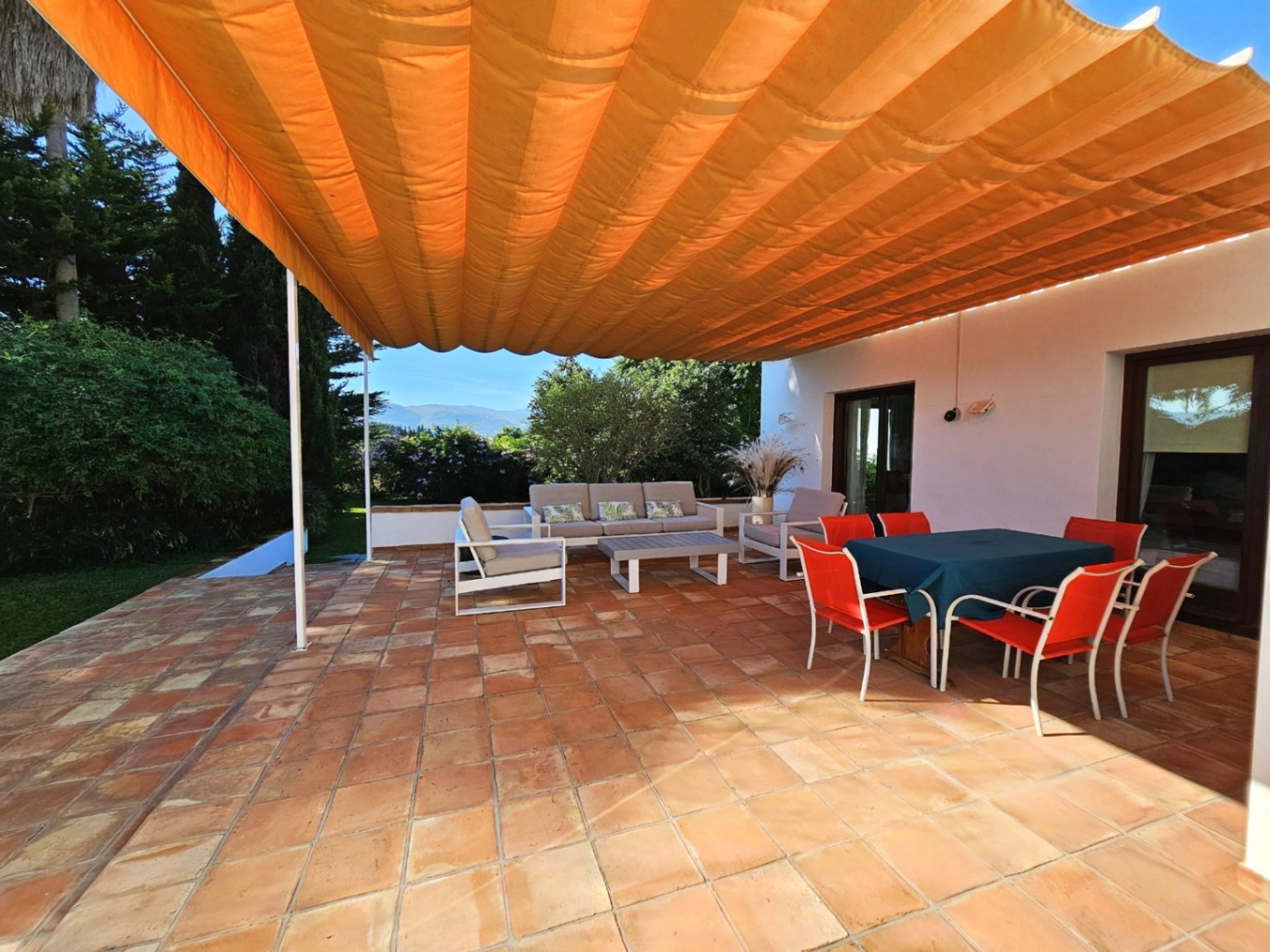Terrasse extérieure avec sols en terre cuite artisanale et un grand store orange. Elle dispose d'un coin repas et d'un salon avec vue sur le jardin.