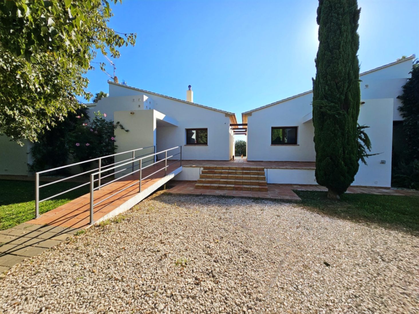 Vue extérieure des deux ailes de la villa avec terrasse en terre cuite, rampe d'accès métallique et allée en gravier bordée d'un cyprès élancé.