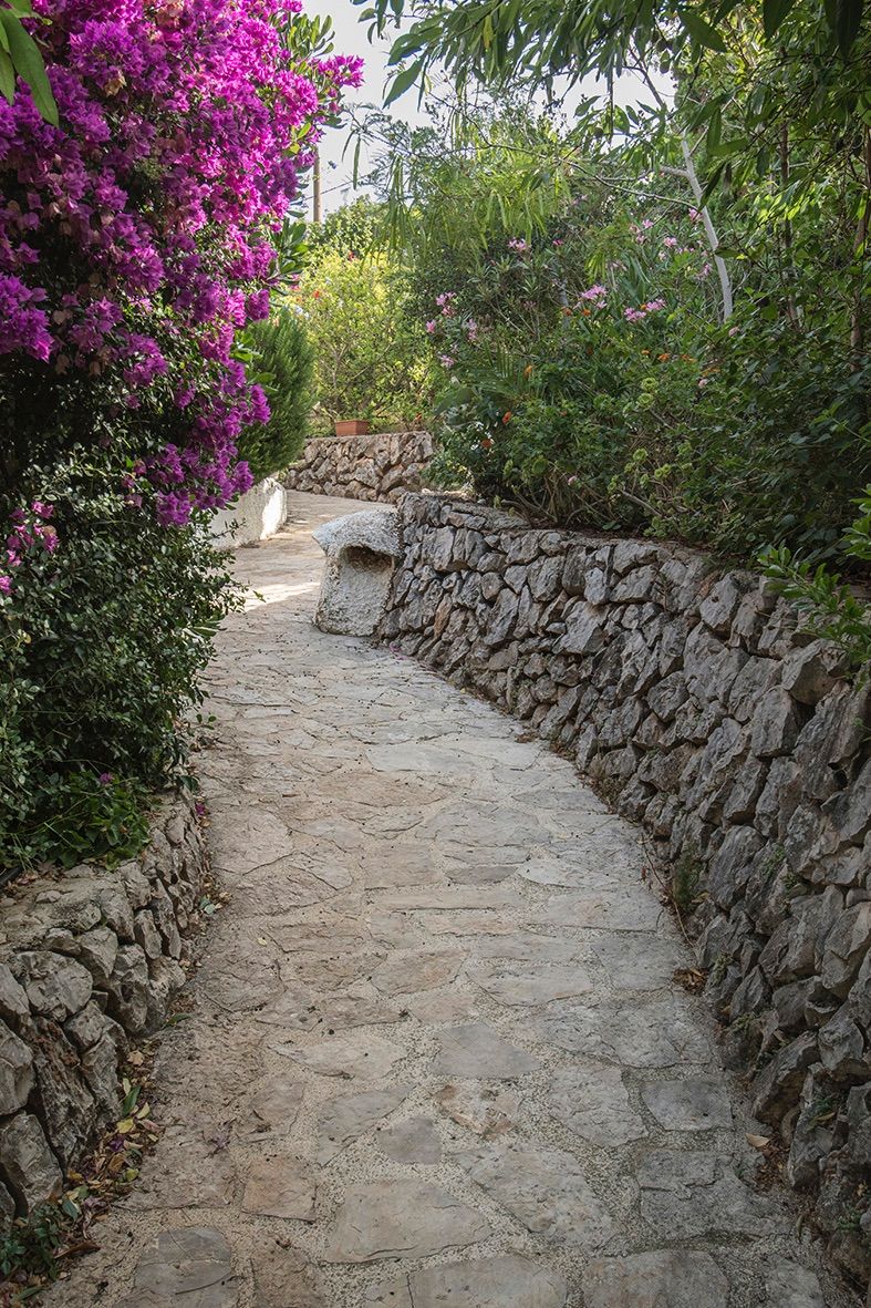 Allée de jardin pavée de pierres irrégulières, bordée de murs en pierre sèche et d'une végétation méditerranéenne luxuriante avec bougainvilliers.
