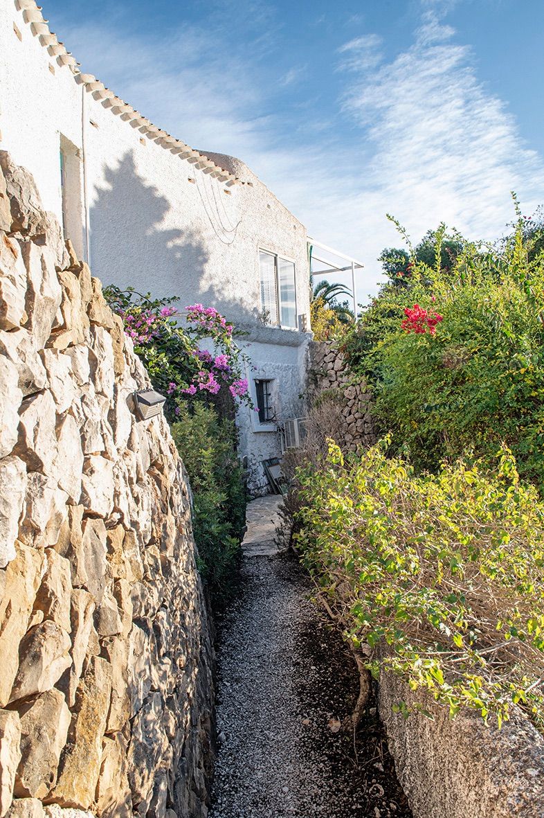 Allée extérieure en gravier bordée de murs en pierre sèche et d'une végétation méditerranéenne luxuriante longeant la façade blanche de la villa traditionnelle.