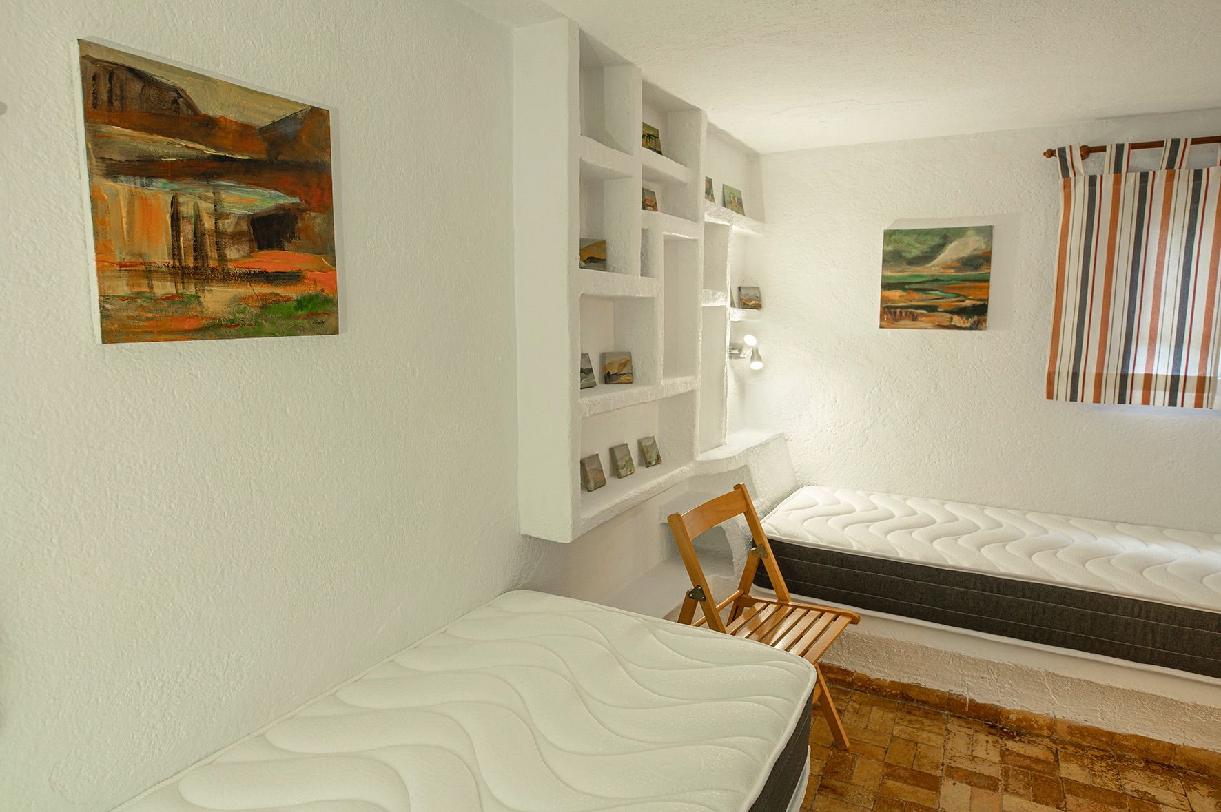 Chambre avec deux lits simples, murs blancs texturés et étagères intégrées sur mesure. Sol en terre cuite traditionnelle et éclairage naturel.