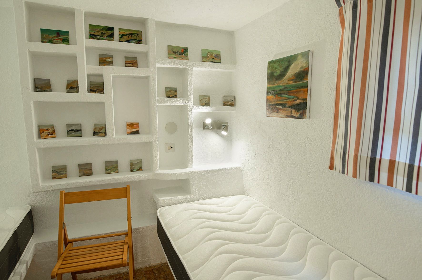 Chambre avec étagères encastrées en maçonnerie dans des murs blancs texturés, agrémentée de petits tableaux paysagers et d'un lit simple.