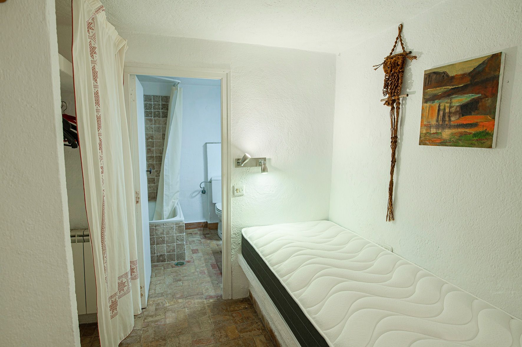 Chambre avec lit simple et salle de bain attenante, carrelage en terre cuite et murs blancs à la finition rustique.