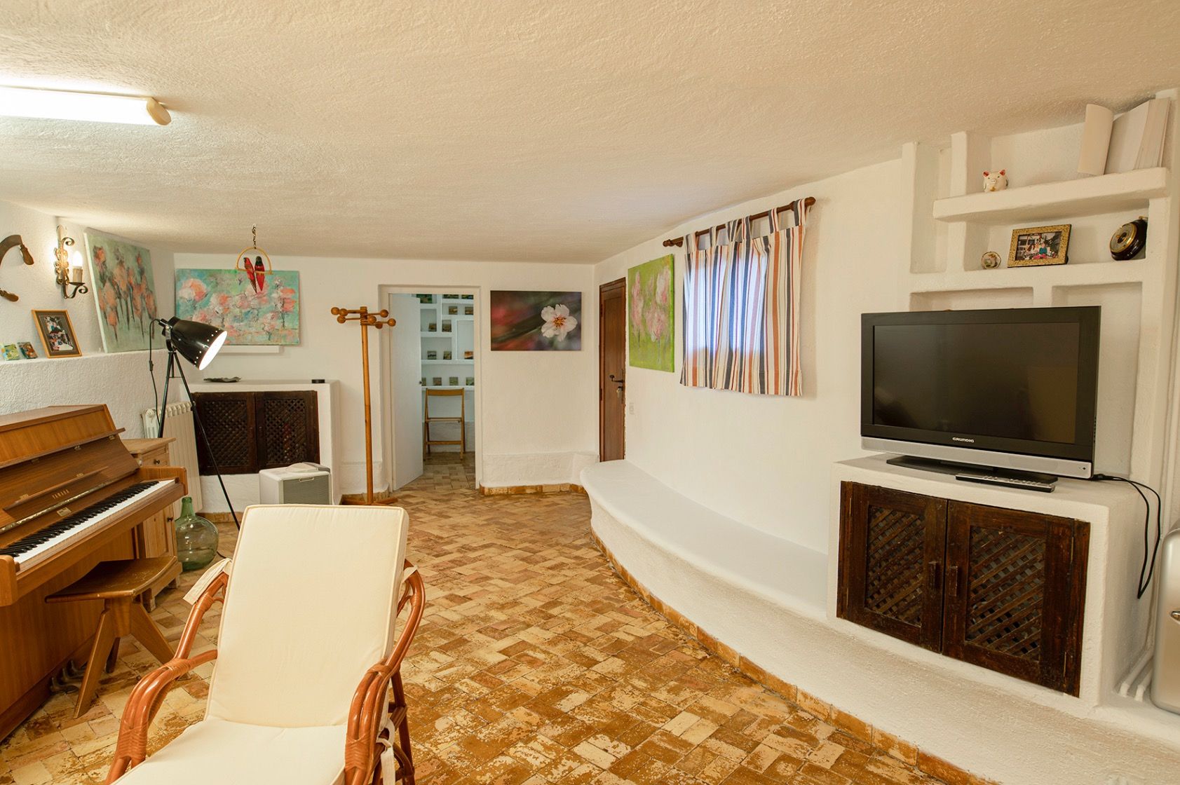 Espace de vie rustique avec sol en terre cuite, piano droit en bois et banquette intégrée en maçonnerie blanche avec rangements à portes en treillis.