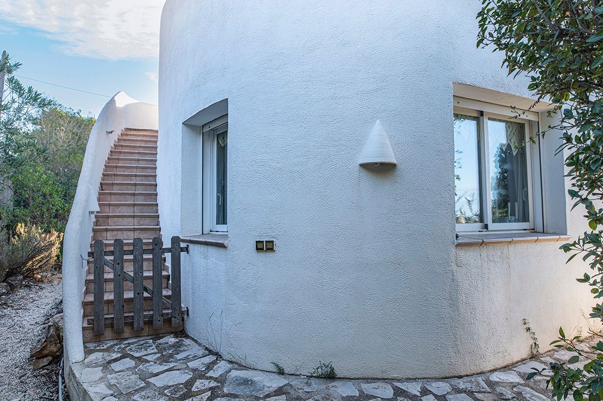 Façade méditerranéenne incurvée avec murs en crépi blanc, escalier extérieur carrelé sécurisé par un portillon en bois et fenêtres à double vitrage.