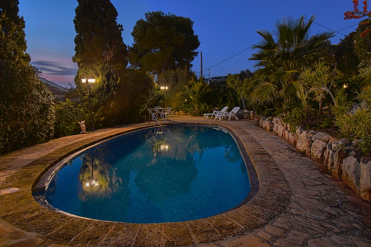 Piscine extérieure éclairée au crépuscule, entourée d'un dallage en pierre naturelle et d'un jardin méditerranéen avec palmiers et cyprès.