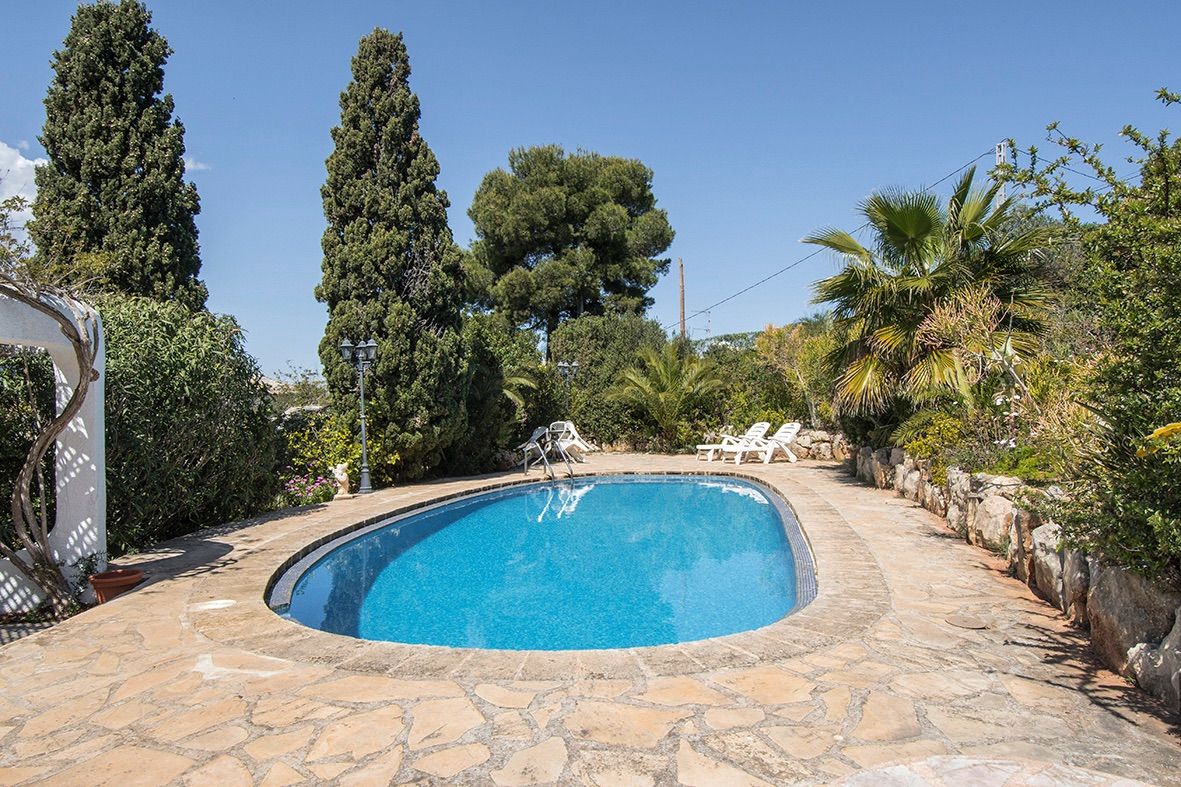Piscine ovale entourée d'une terrasse en pierre naturelle et d'un jardin méditerranéen arboré de cyprès et de palmiers.