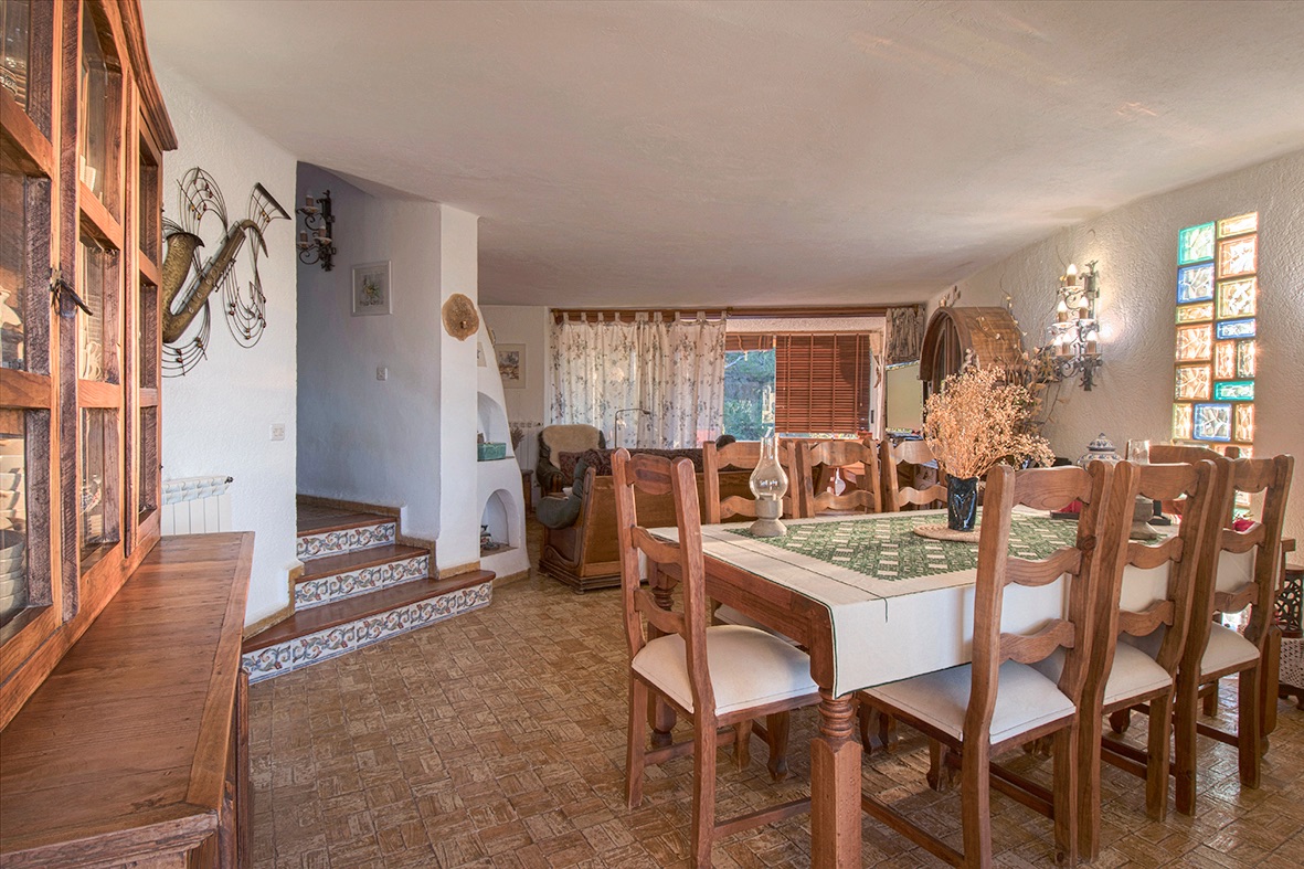 Salle à manger rustique avec mobilier en bois, sol en grès et marches ornées d'azulejos. Un mur en briques de verre colorées apporte une lumière naturelle diffuse.