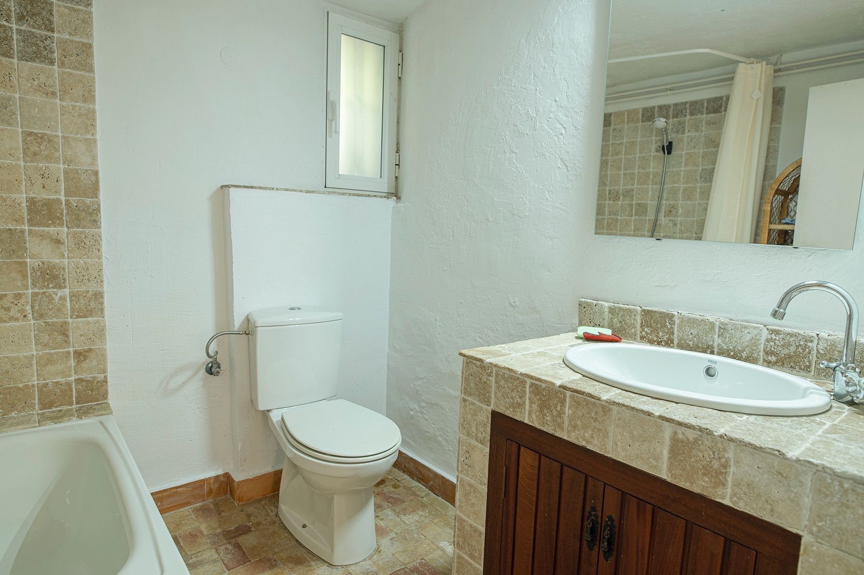 Salle de bain de style rustique avec plan vasque en pierre, rangements en bois, baignoire blanche et murs en plâtre avec fenêtre oscillo-battante.