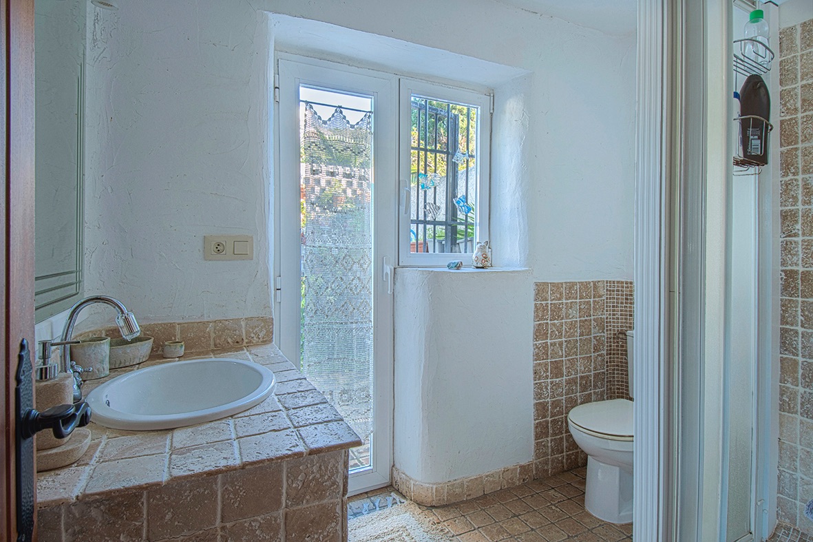 Salle de bain rustique avec plan de toilette carrelé, vasque encastrée et murs blancs texturés, complétée par une porte vitrée apportant une luminosité naturelle.