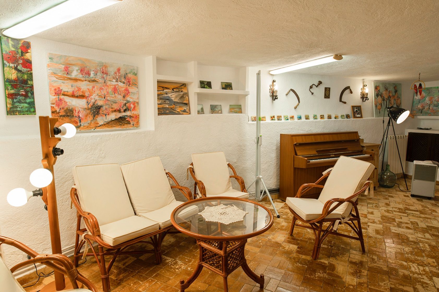 Salon avec mobilier en bambou, sol en terre cuite et murs blancs texturés. L'espace comprend un piano droit, des niches artistiques et une table basse en verre.