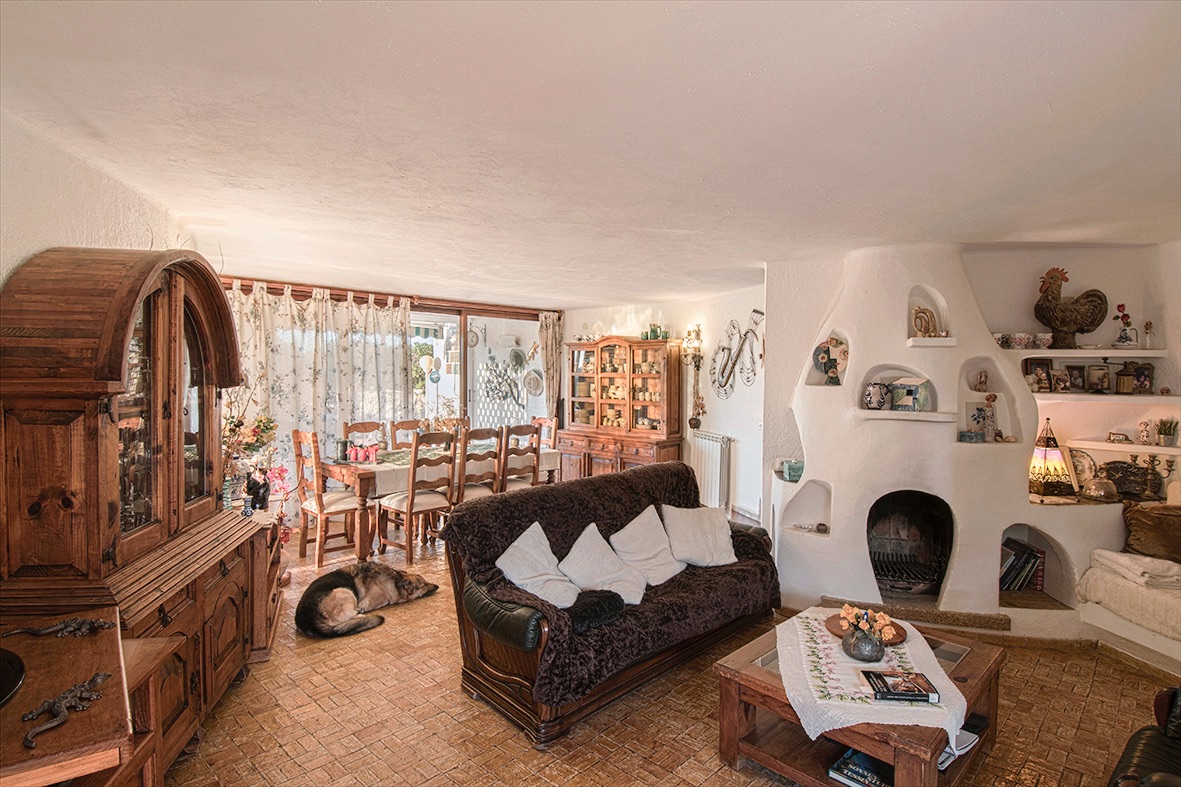 Salon-salle à manger rustique avec une cheminée sculptée blanche unique, des meubles en bois massif et un sol en terre cuite traditionnelle sous une lumière naturelle.