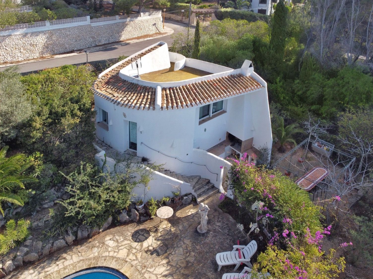 Vue aérienne de la villa secondaire avec murs blancs incurvés, espace barbecue intégré, escaliers en pierre et terrasse pavée entourée de jardins méditerranéens.