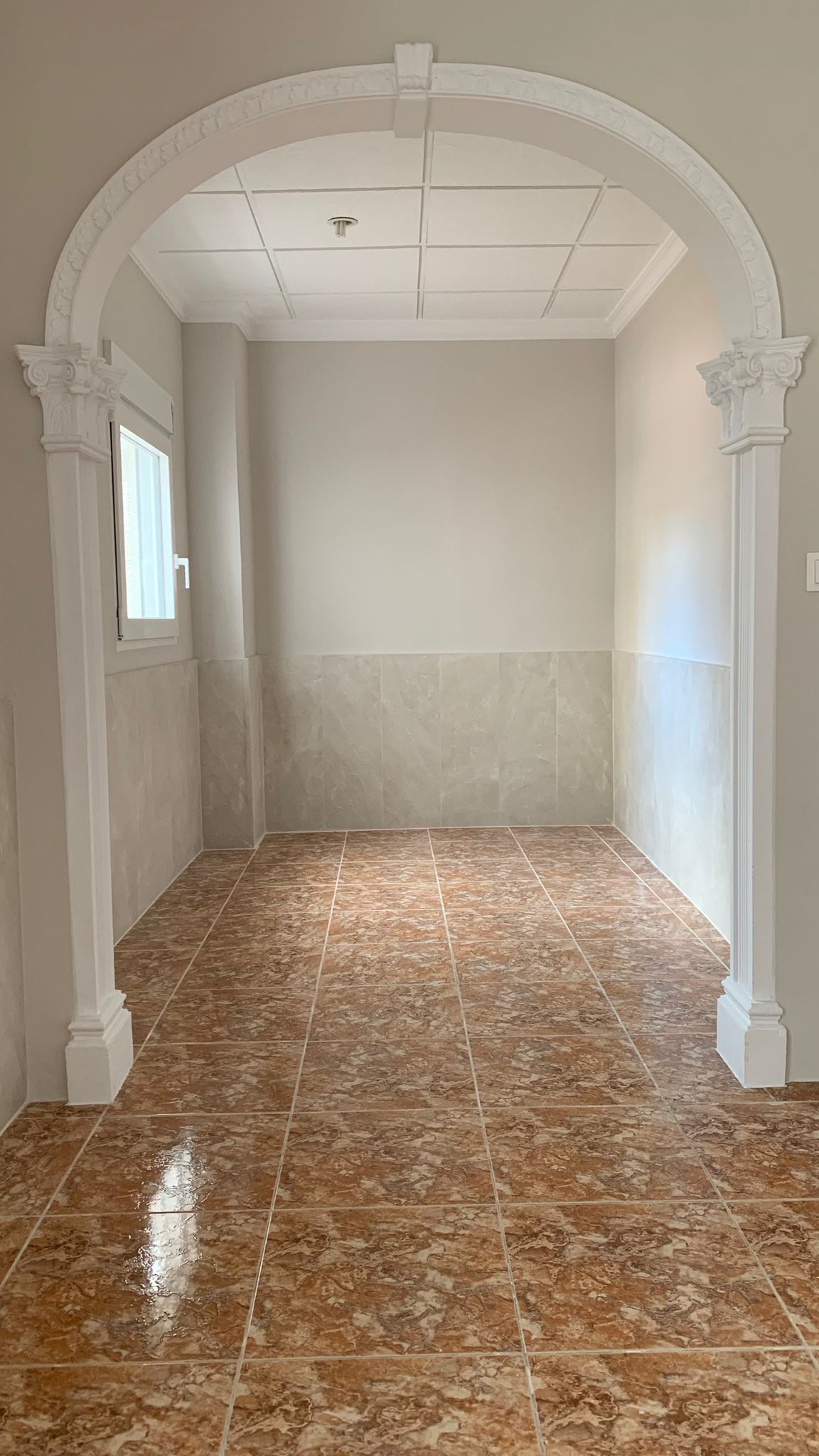 Hall d'entrée avec arche décorative classique, sol en grès cérame aux tons terreux et soubassement en carrelage gris. Une fenêtre latérale apporte de la clarté naturelle.