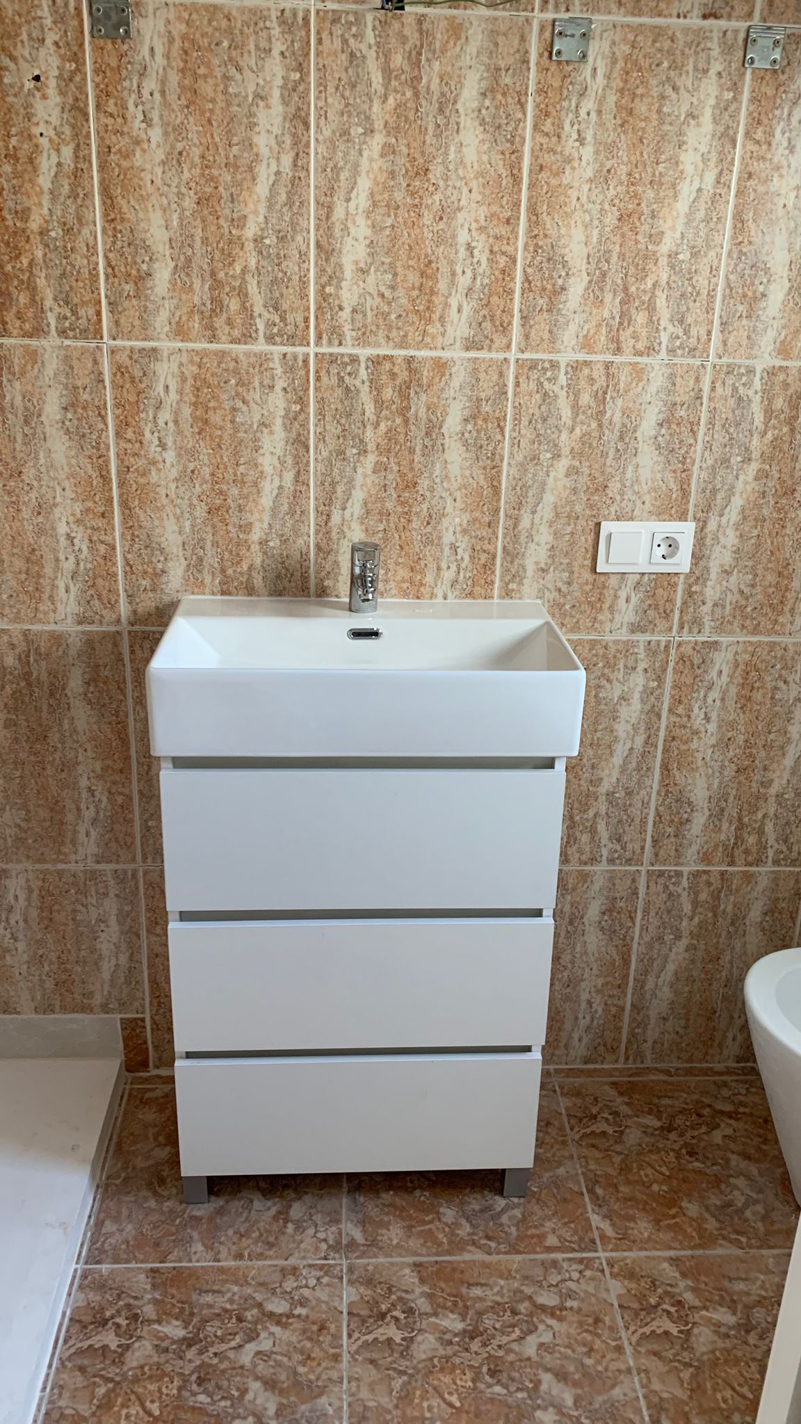 Meuble vasque moderne blanc à trois tiroirs avec évier intégré, entouré de carrelage mural et de sol aux tons terreux effet marbre.