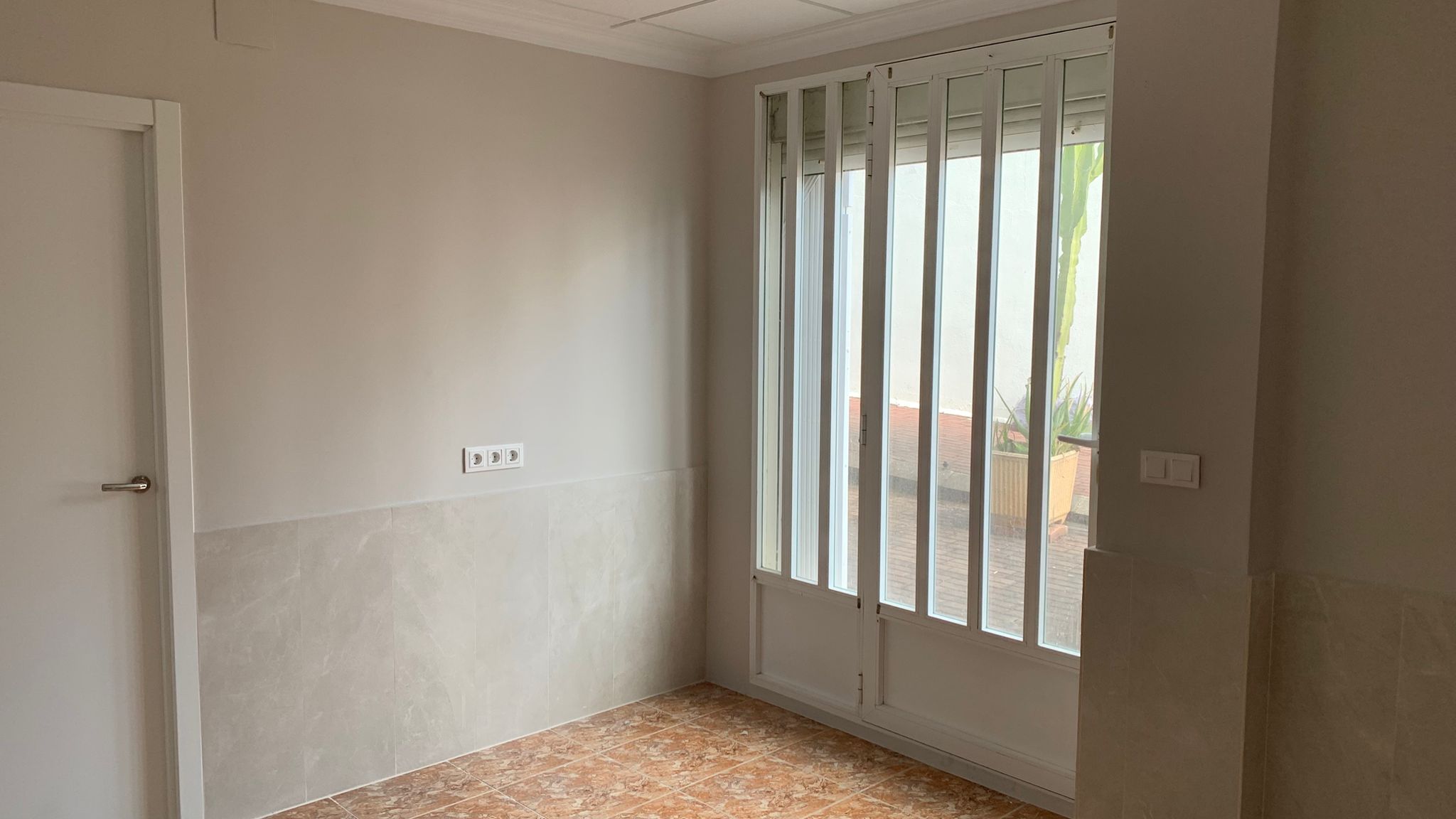 Pièce avec sol en grès, murs partiellement carrelés et porte vitrée avec grilles de sécurité blanches offrant un accès direct à l'extérieur.