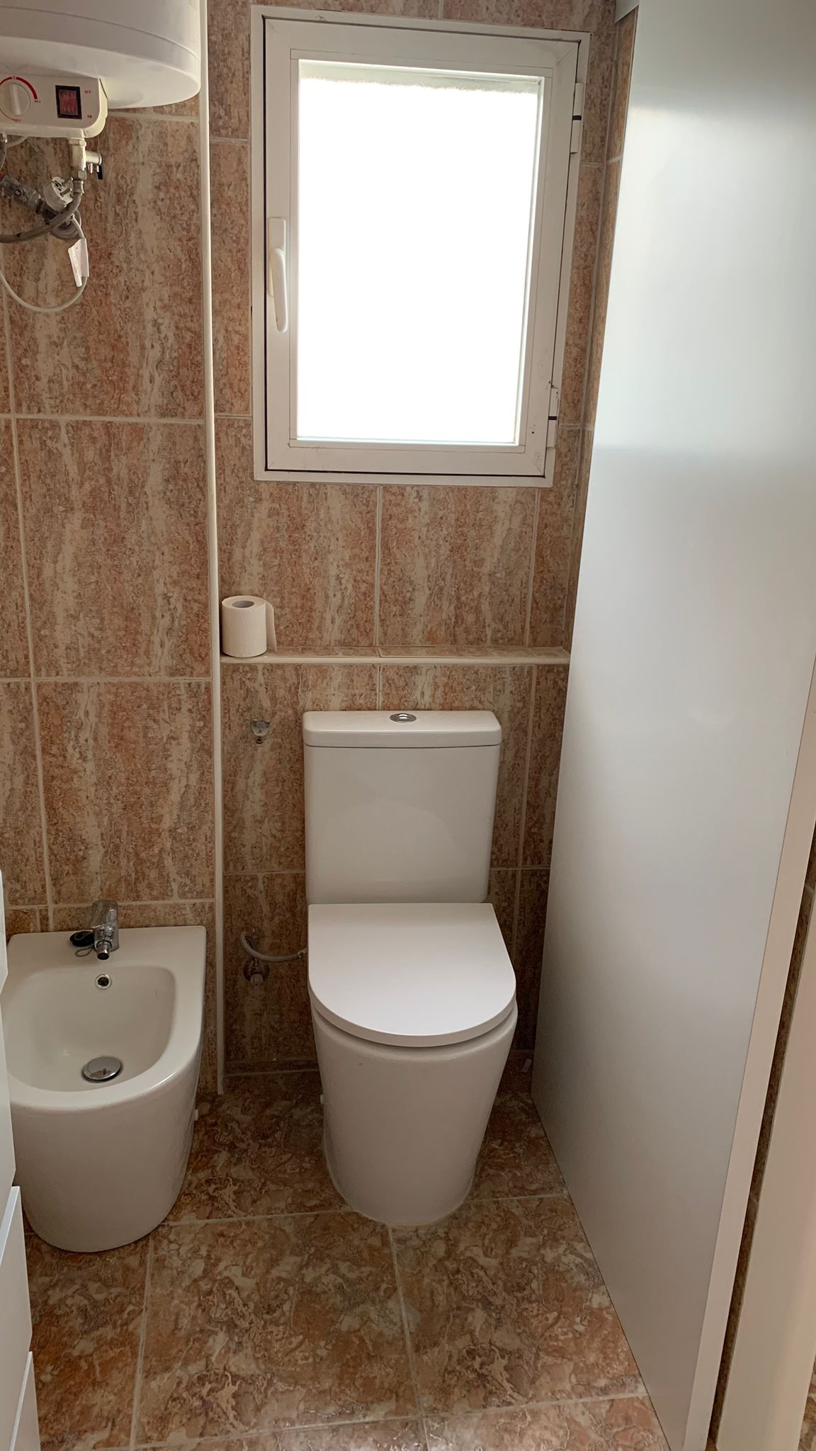 Salle de bains avec toilettes et bidet en porcelaine, carrelage aux tons terreux, paroi de douche en verre translucide et fenêtre.