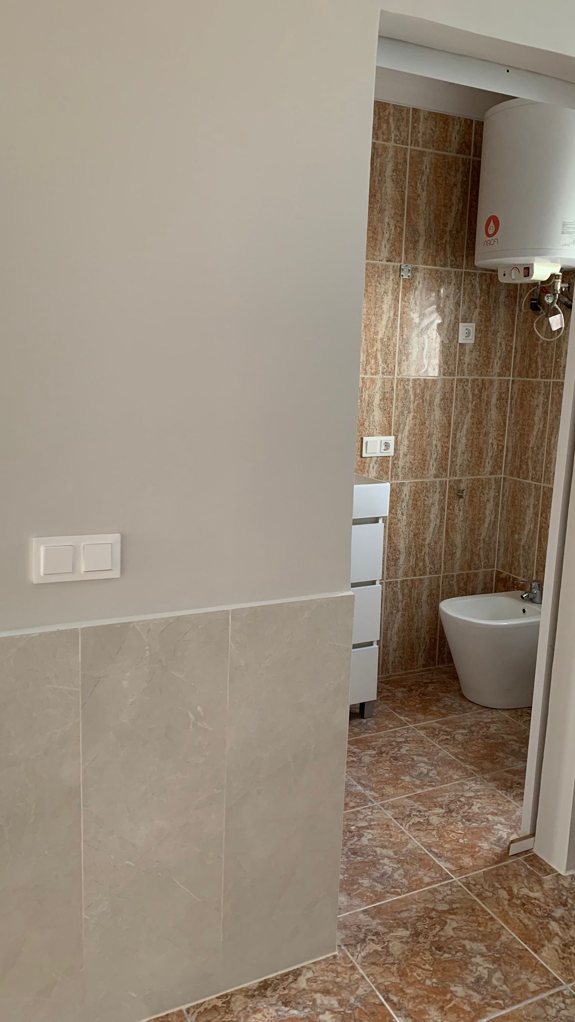 Vue de la salle de bain avec bidet, chauffe-eau électrique et carrelage aux tons terreux, depuis un couloir avec soubassement en marbre.
